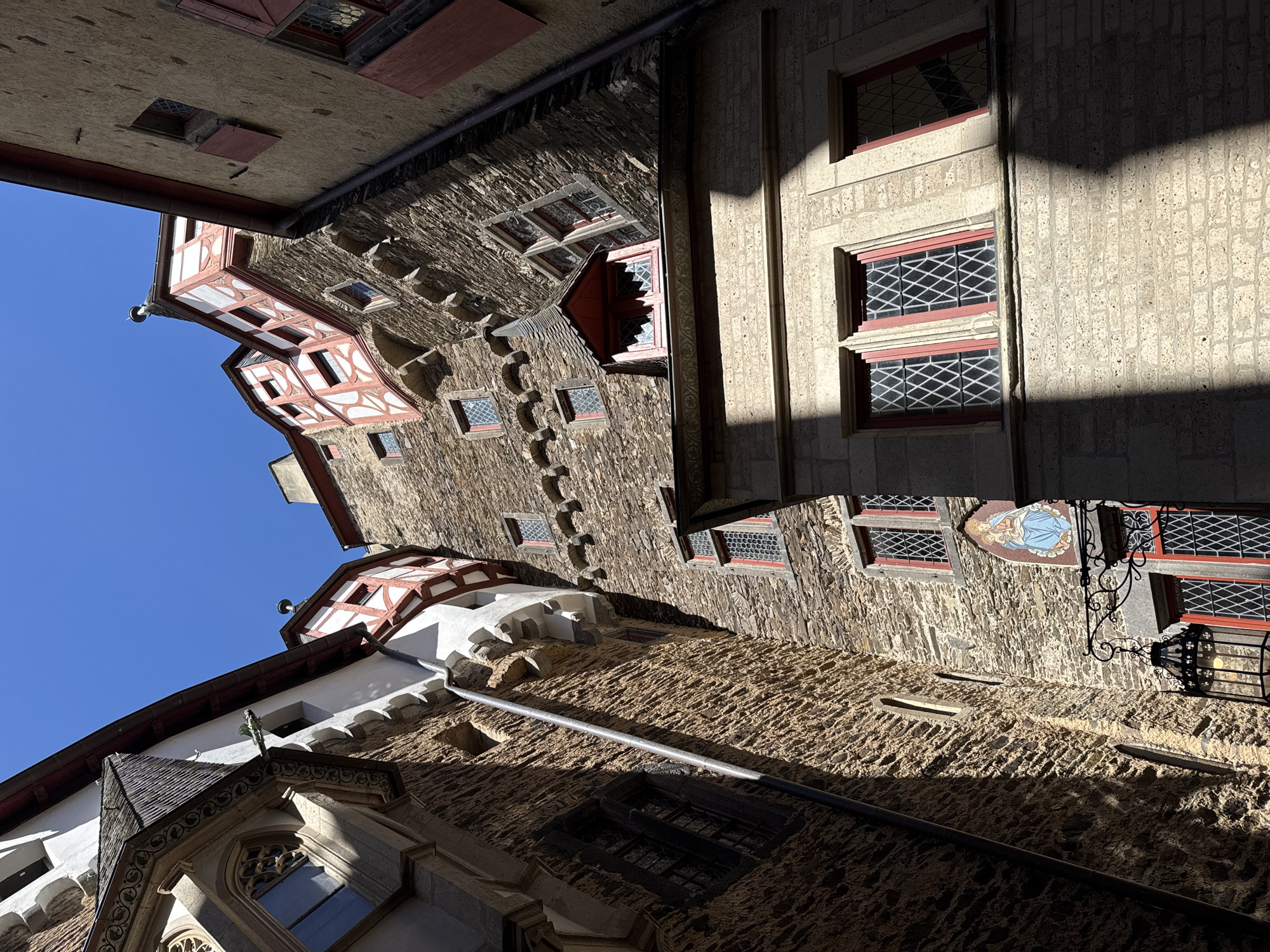 The image depicts the architecture of Eltz Castle in Wierschem, Germany, captured from a low angle looking upwards. The castle walls are constructed of stone and masonry, showcasing a variety of architectural styles with features such as windows, turrets, and decorative elements. The sky is visible in the upper left corner, providing a backdrop of clear blue. A mix of sunlight and shadows plays across the surfaces of the castle, highlighting the textures and depth of the structure.