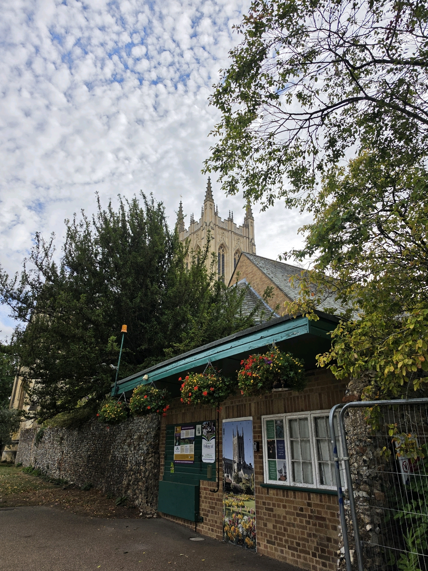 The image shows a picturesque scene in West Suffolk, United Kingdom. In the foreground, a small brick building with a teal-colored overhang is visible. Hanging baskets filled with red and yellow flowers adorn the overhang. A large stone wall runs along the left side of the building, and a paved pathway is in front of it.
Behind the building, lush green trees partially obscure a majestic Gothic-style church with spires and intricate stonework. The church's architecture suggests it is a historical landmark, possibly a cathedral or a large parish church. The sky above is a mix of blue and white clouds, creating a dynamic backdrop. A metal fence is present on the right side of the image, suggesting a boundary or construction area. The overall impression is one of natural beauty combined with historical architecture.