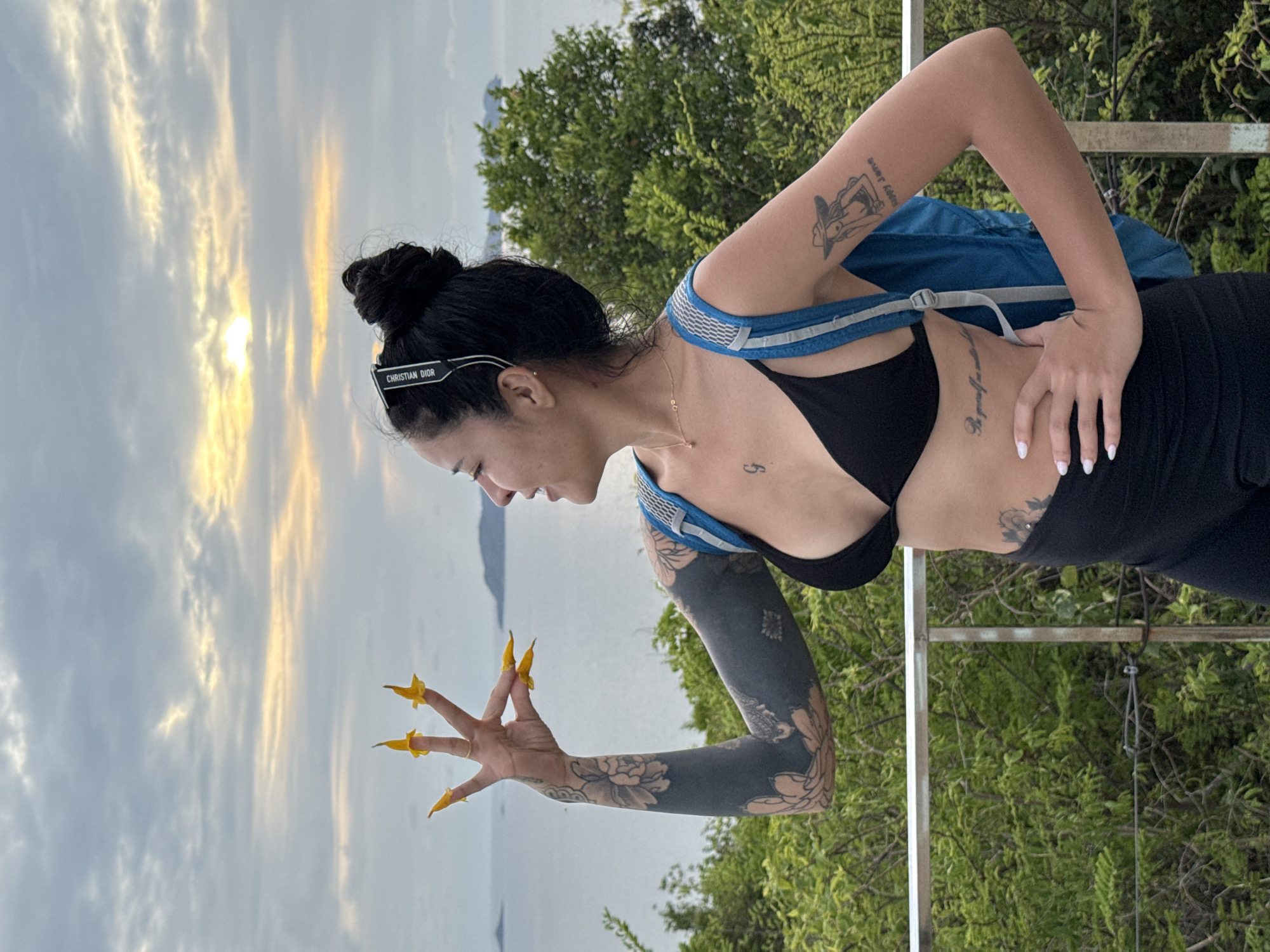 A woman stands with her back to greenery and faces a vista of water and a sunset-streaked sky. She wears a black bikini top and black leggings. A blue backpack rests on her shoulders. Her hair is pulled into a bun, and she wears sunglasses atop her head. Her right arm is outstretched, fingers spread, with bright yellow floral-shaped decorations attached to the nails. Her left hand rests on her torso. Extensive tattoos cover her left arm. A metal railing runs vertically along the right side of the image. The scene takes place in Pattaya City, Thailand.