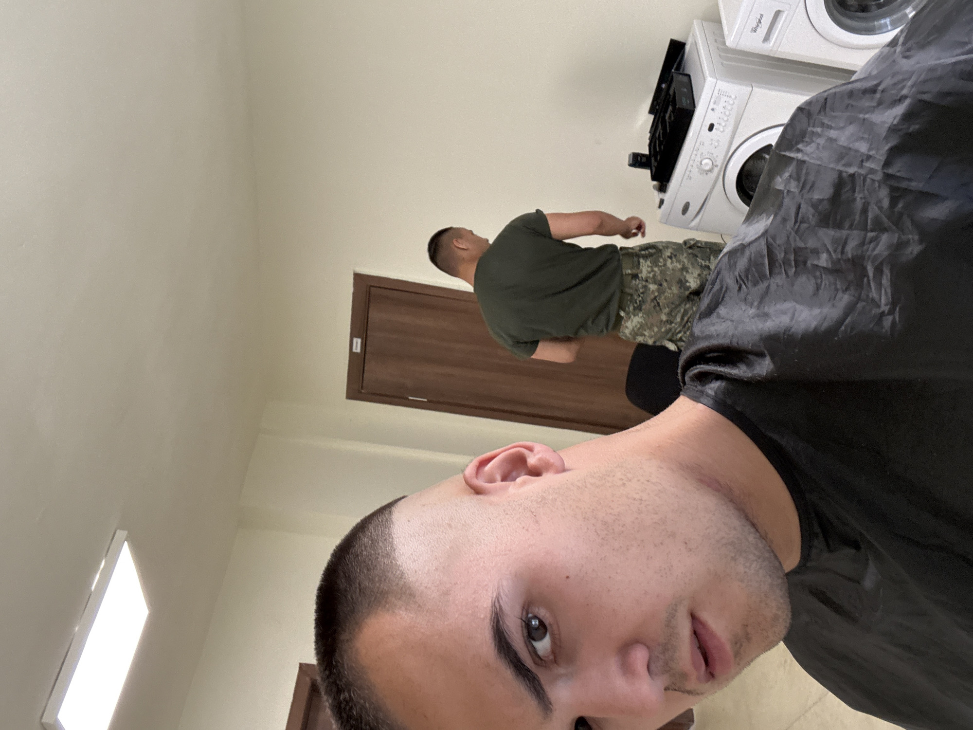A selfie is taken from a low angle, showing the face of a man with a buzz cut and a black cape draped over his shoulders. He has dark hair, brown eyes, and a light beard. In the background, another man in a green t-shirt and camouflage pants is standing next to a white washing machine. The scene appears to be indoors, possibly in a laundry room or a barber shop, with beige walls and a bright fluorescent light fixture on the ceiling. There are two washing machines visible, one white and one with a Whirlpool logo. A dark-colored box with black items is on top of the white washing machine. A wooden door is also visible in the background.