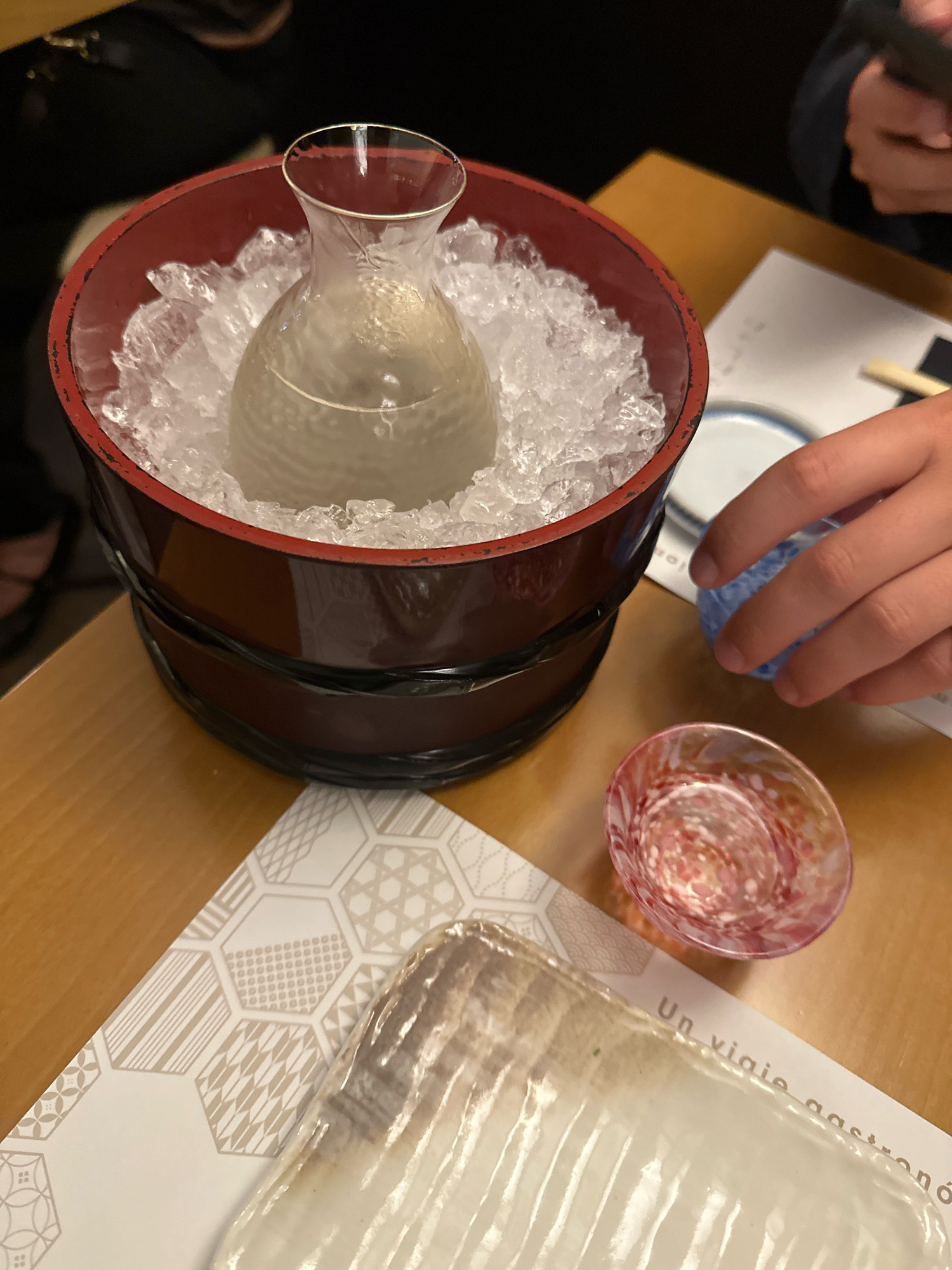 A close-up overhead shot captures a dining scene with a sake bottle nestled in a wooden ice bucket on a wooden table. The clear glass bottle, containing a pale liquid, is surrounded by crushed ice, and a subtle pattern is visible on its surface. To the right, a hand is seen reaching for a small blue sake cup, while another ornate pink and red glass cup rests nearby. A patterned paper placemat with hexagonal designs and text reading "Un viaje gastronó" is partially visible in the foreground, suggesting a Spanish dining experience, likely in Madrid, Spain, given the implied context. The lighting is warm, casting soft shadows on the table.
