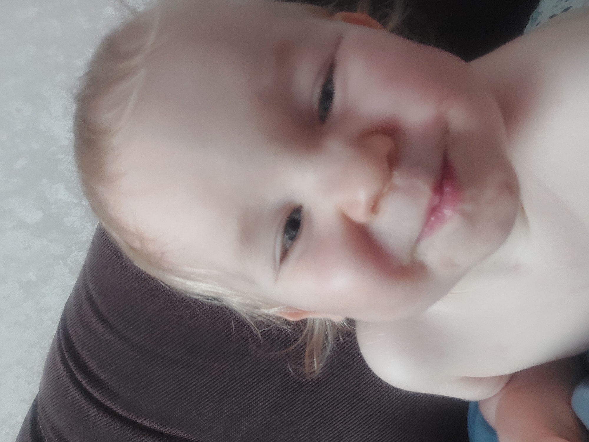 A close-up photograph shows a baby lying on a dark brown fabric surface. The baby has fair skin, blond hair, and light-colored eyes. The baby's face is slightly tilted towards the viewer. The baby's expression is gentle and lips are slightly parted. The background is out of focus and appears to be a light grey wall. There is no discernible evidence connecting the photo to the city of Chaikovsky, Russia.