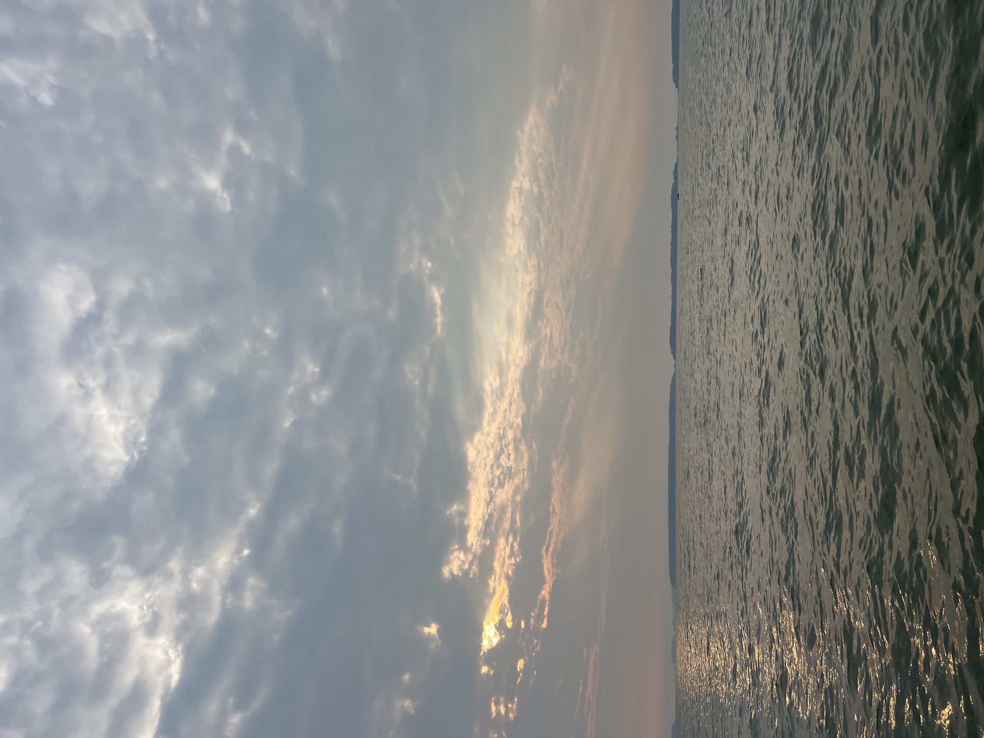 The image depicts a wide body of water under a cloudy sky. The sun is partially obscured by clouds, allowing rays of light to break through and illuminate the water's surface, creating a shimmering effect. In the distance, a thin, dark line of land with trees is visible on the horizon. There is a small boat on the water. The overall mood is serene and somewhat overcast, with the light suggesting either dawn or dusk. This scene is not in Novopichugovo, Russia; it is a generic landscape.