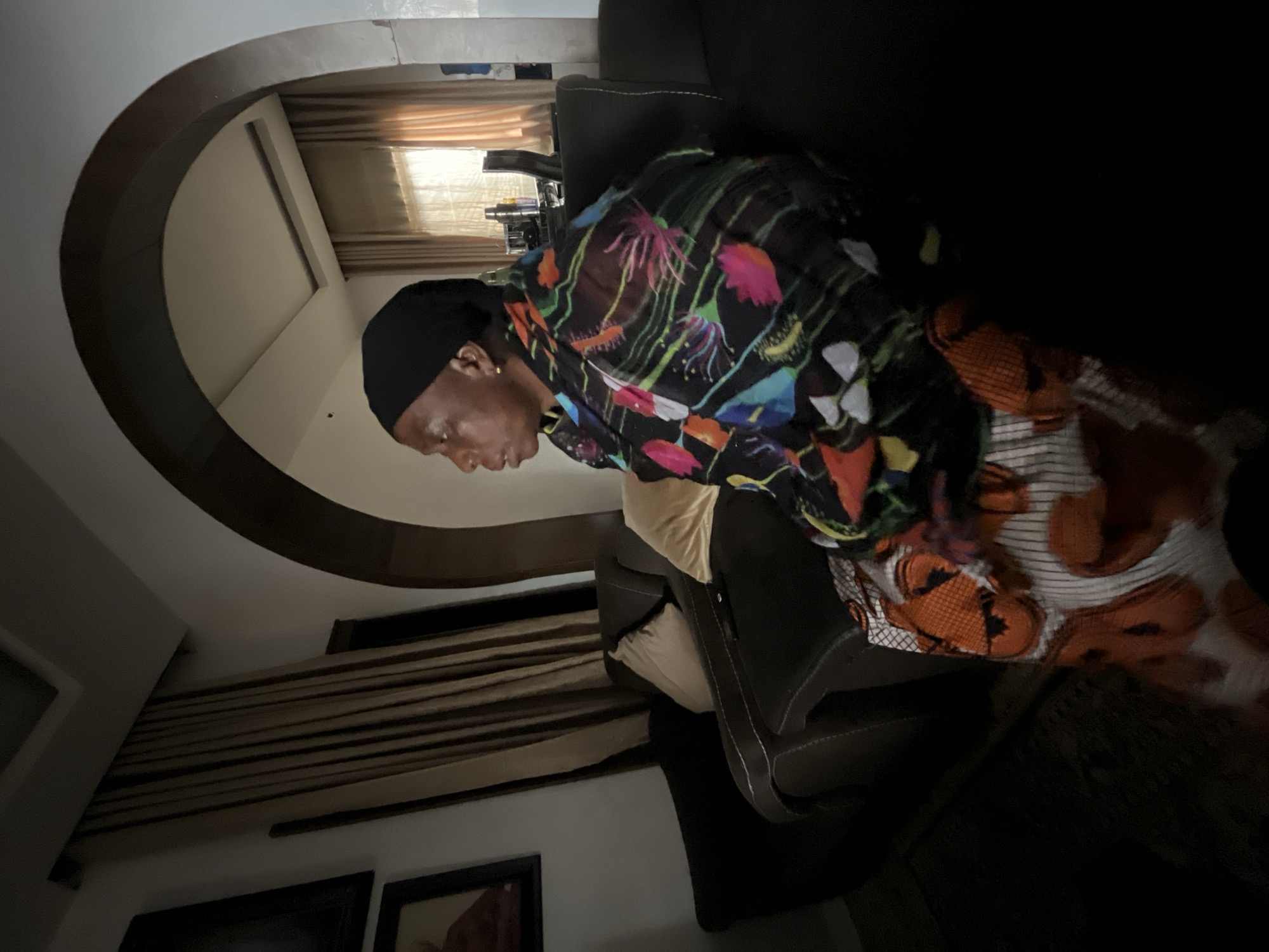 A person with dark skin and a black head covering sits on a dark-colored chair in what appears to be an indoor setting. The person is wearing a colorful, patterned top and orange and white patterned bottoms. The environment includes an arched opening, a window with closed blinds, and framed pictures on a wall. There is no explicit visual information to determine that this image was taken in Jos, Nigeria.