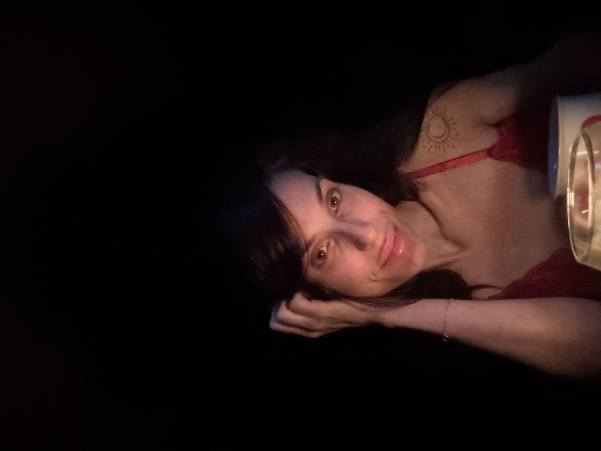 The image shows a woman lying down in a very dark, low-light setting, likely at night or in a dimly lit room. She is positioned with her head resting on her hands, looking directly at the camera with a calm, slightly smiling expression. She has dark hair and a fair complexion.
She is wearing a red top, possibly lingerie, with a thin strap visible over her left shoulder. A distinct circular tattoo, featuring a sun with rays, is visible on her left shoulder. A delicate chain bracelet adorns her left wrist. On the far right of the frame, a portion of a glass or container, possibly holding a candle or reflecting light, is visible and appears to be the primary light source illuminating her face and upper body. The vast majority of the background is in complete darkness, creating a stark contrast and intimate atmosphere. No text is visible in the image. The scene is set in Pleven, Bulgaria.