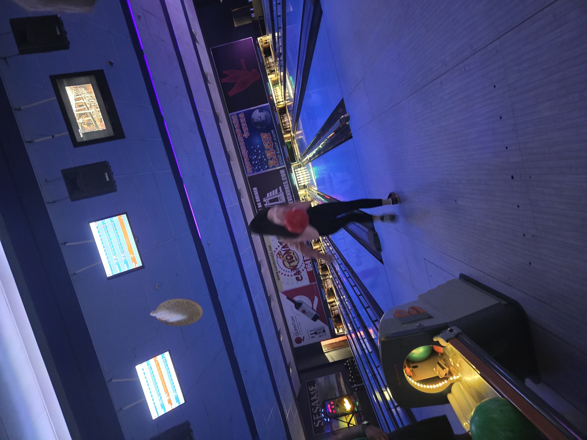 A bowling alley in Pleven, Bulgaria is depicted with a woman in black pants and a black top, preparing to bowl with a red bowling ball. The alley is illuminated with blue and purple lighting. Monitors display scores and advertisements. Bowling ball return mechanisms are visible along the alley, with bowling balls ready for use. Banners and signage, including one for "Sesame," adorn the walls.
