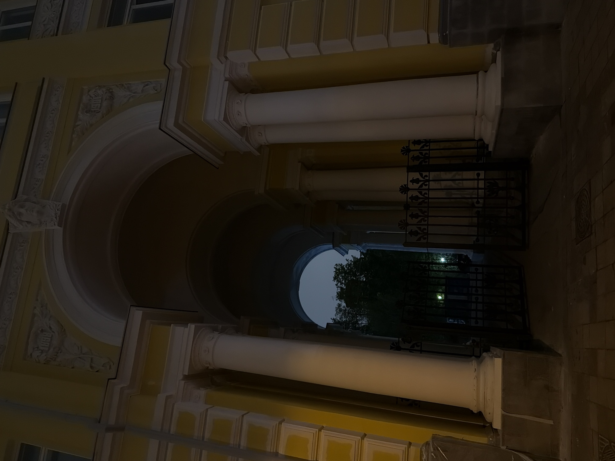 The image shows a view through an archway into a courtyard or garden in Moscow, Russia. The archway is part of a building with classical architectural elements including columns and decorative moldings. A wrought iron gate is visible at the end of the archway, beyond which are trees and dimly lit grass. The building is painted yellow, the columns white, and the wrought iron is black. The scene appears to be at night or during twilight, with low levels of illumination.