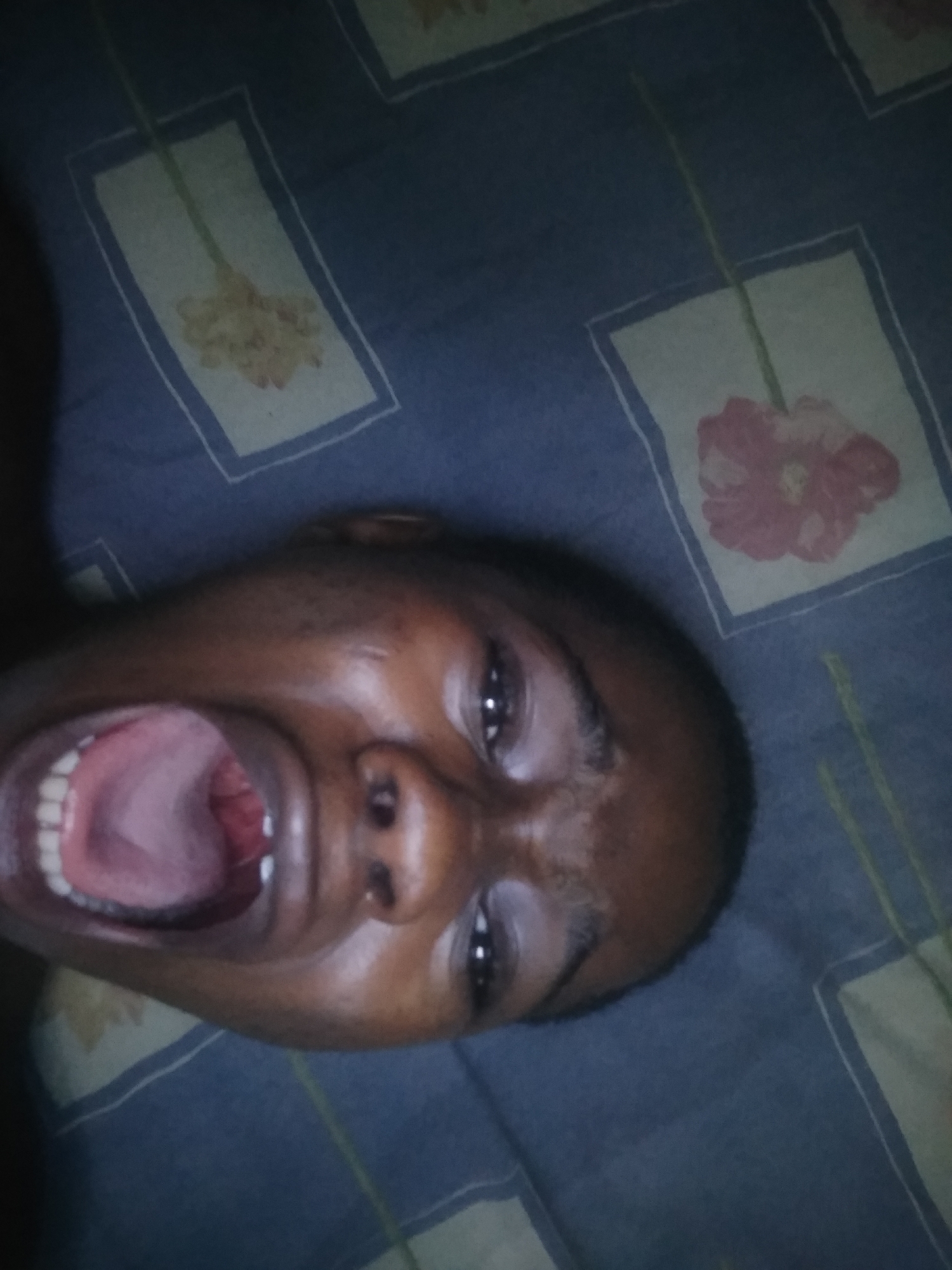 A person is lying on a bed with a floral-patterned bedsheet, looking up at the camera. The person's mouth is open wide. The location is unknown as Shangisha is not included in the data.