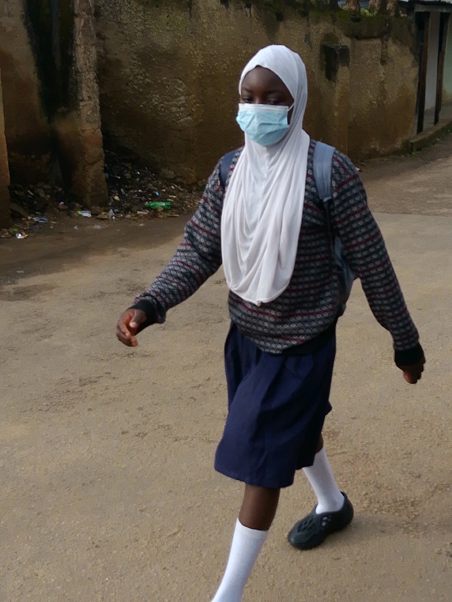 A young person wearing a white hijab, a blue face mask, a patterned sweater, and a dark blue skirt walks on a dirt road. The person is carrying a backpack. In the background, there is a textured wall with some debris on the ground. The environment appears to be an outdoor urban setting in Jos, Nigeria.