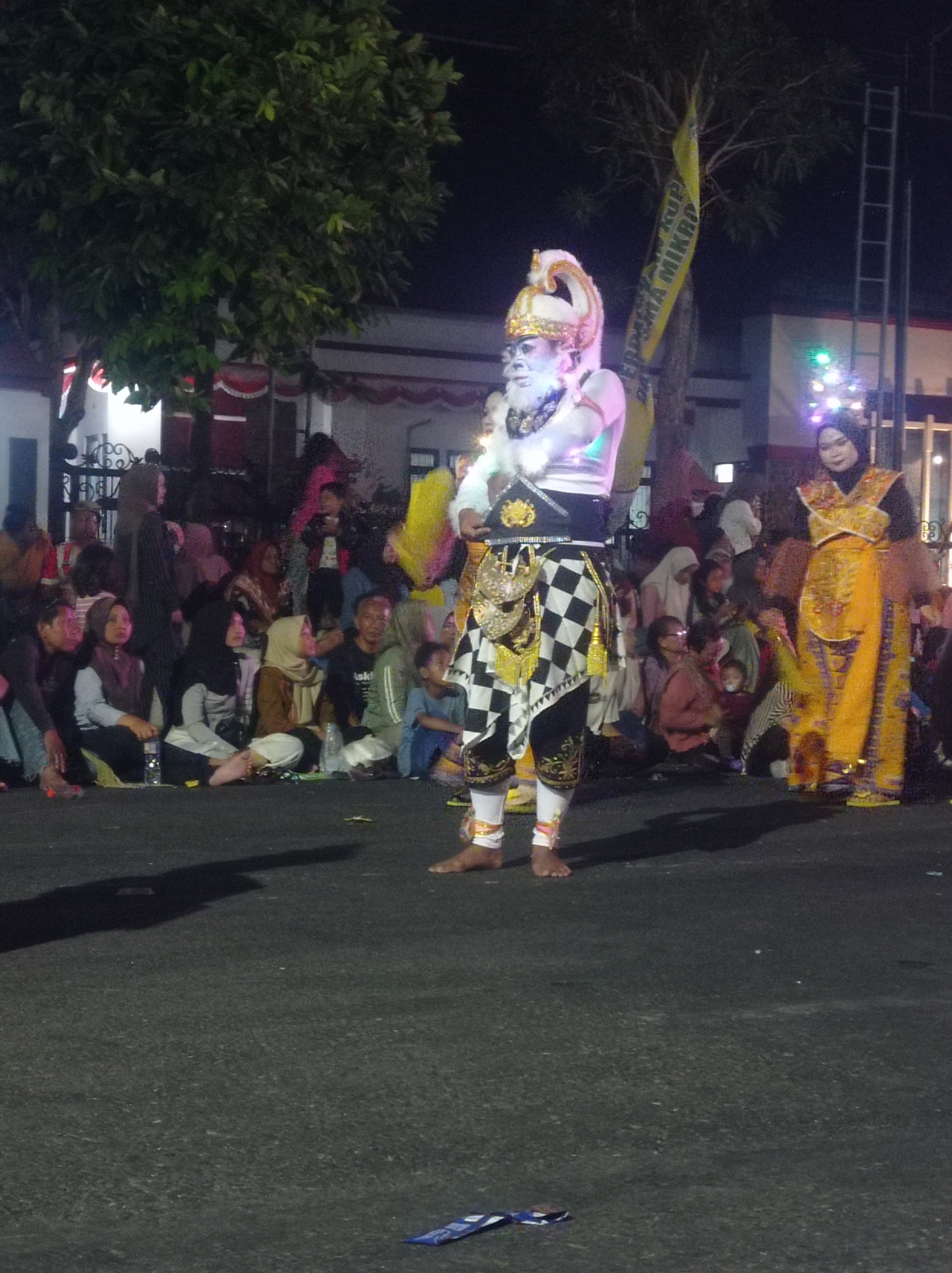 A medium shot captures a traditional performance at night in Pandean, Indonesia. The central figure, a performer with white face paint, a white beard, and an elaborate golden headdress, stands center, adorned in a costume featuring a black and white checkered sarong and a white top with gold embellishments. To the right, another performer in a yellow and gold traditional outfit stands facing forward. A crowd of spectators, dressed in everyday clothing and headscarves, sits on the ground in the background, watching the performance. Trees and a building with red and white banners are visible behind the spectators. The ground is asphalt. The lighting suggests a festive outdoor event.