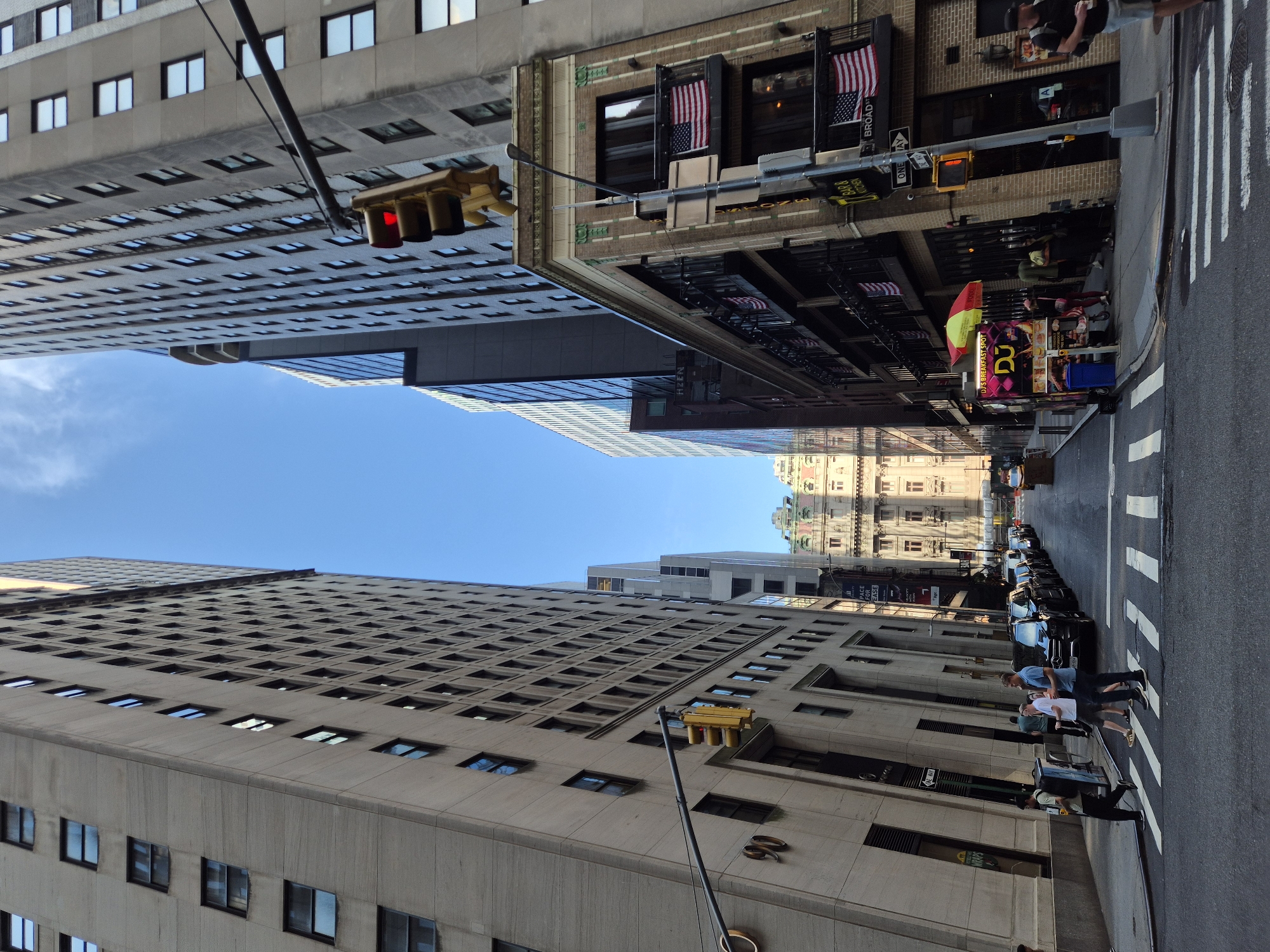 The image captures a street scene in the financial district of New York City, United States of America. The perspective is from a street level looking down a canyon of tall buildings on a clear day with a bright blue sky. On the left, a light-colored, textured concrete building with numerous windows dominates the view. The building has the number "90" clearly visible on its facade. On the right, a brick building with balconies adorned with American flags stands out, with a sign that reads "BROAD ST" and another sign for "DJ'S BREAKFAST SPOT" above a food cart. Further down the street, more modern glass-faced buildings rise, and in the distance, an ornate, older building with a light-colored facade and a greenish roof is visible.
Several cars are parked along the right side of the street, and a crosswalk with white stripes is present in the foreground. A few pedestrians are walking across the street and on the sidewalk. One man in a blue shirt and another in a white t-shirt are crossing the street from left to right. Another individual is walking on the sidewalk with a backpack. A traffic light is visible, currently showing red. A street sign indicating "ONE WAY" is also present. The overall impression is of a bustling urban environment.