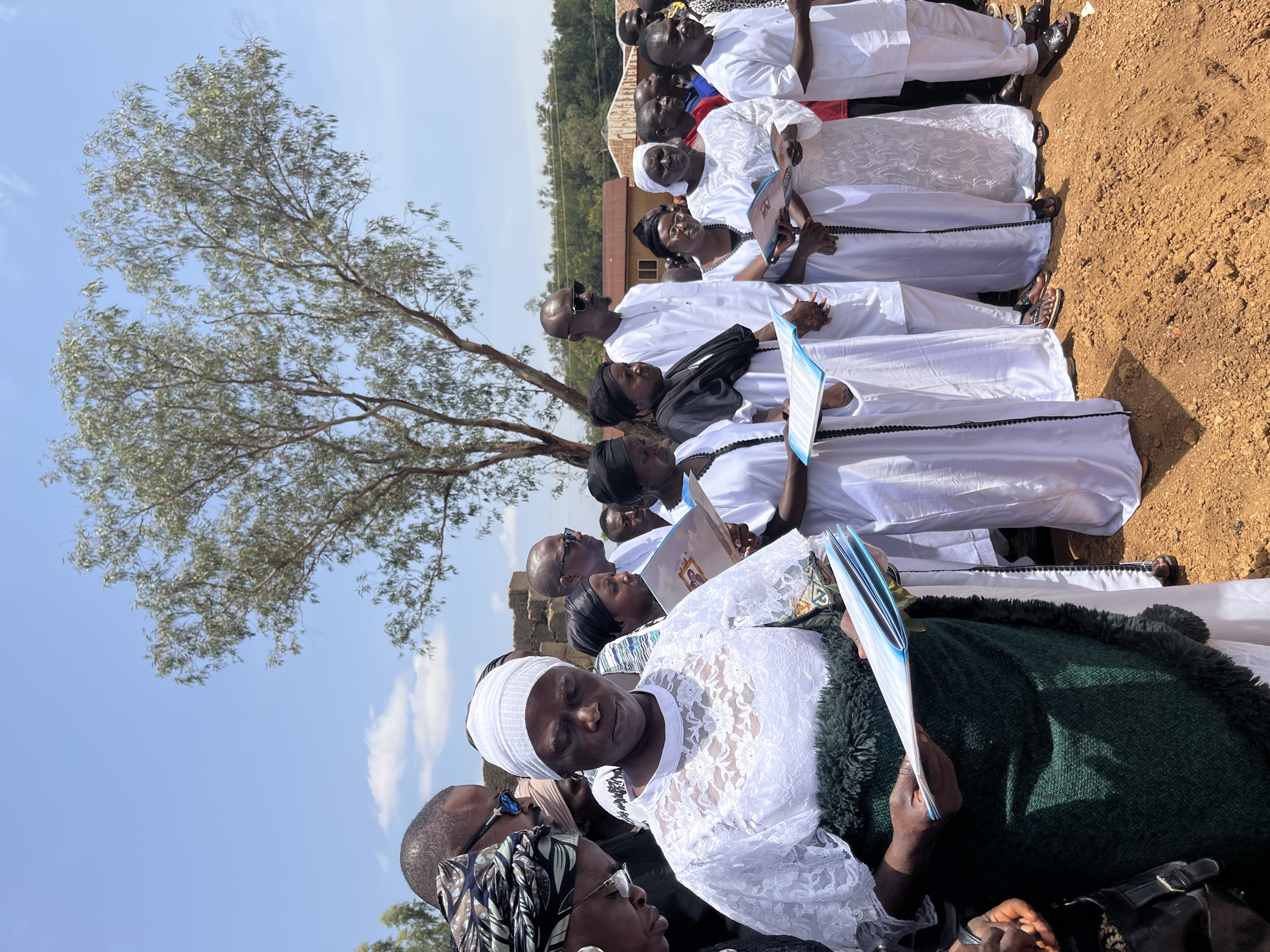 A group of people, predominantly Black, stand outside on a sunny day, possibly in Dogon Dinia, Nigeria. Most of them are dressed in white, some with white head coverings or lace detailing. They are holding booklets or programs in their hands. A tree with thin, light-colored leaves is prominent on the left side of the frame. The ground appears to be reddish-brown earth. Some people are wearing dark sunglasses.
