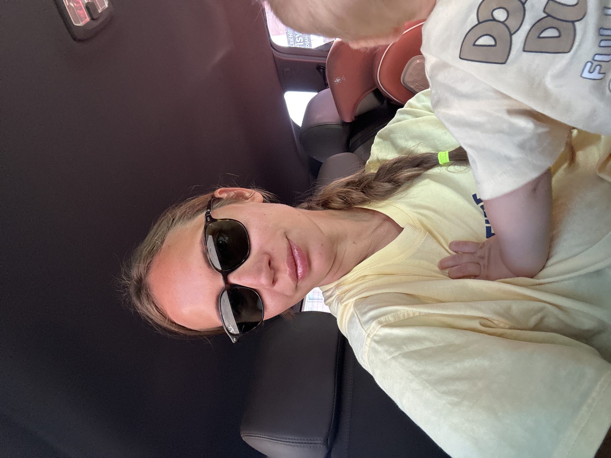 A woman wearing sunglasses and a yellow shirt is seated in a car. A baby wearing a light yellow shirt is nestled on her chest. The car appears to be parked, given the interior view with the roof and front seats visible. The photograph was likely taken in Fethiye, Turkey, based on the surrounding image metadata.