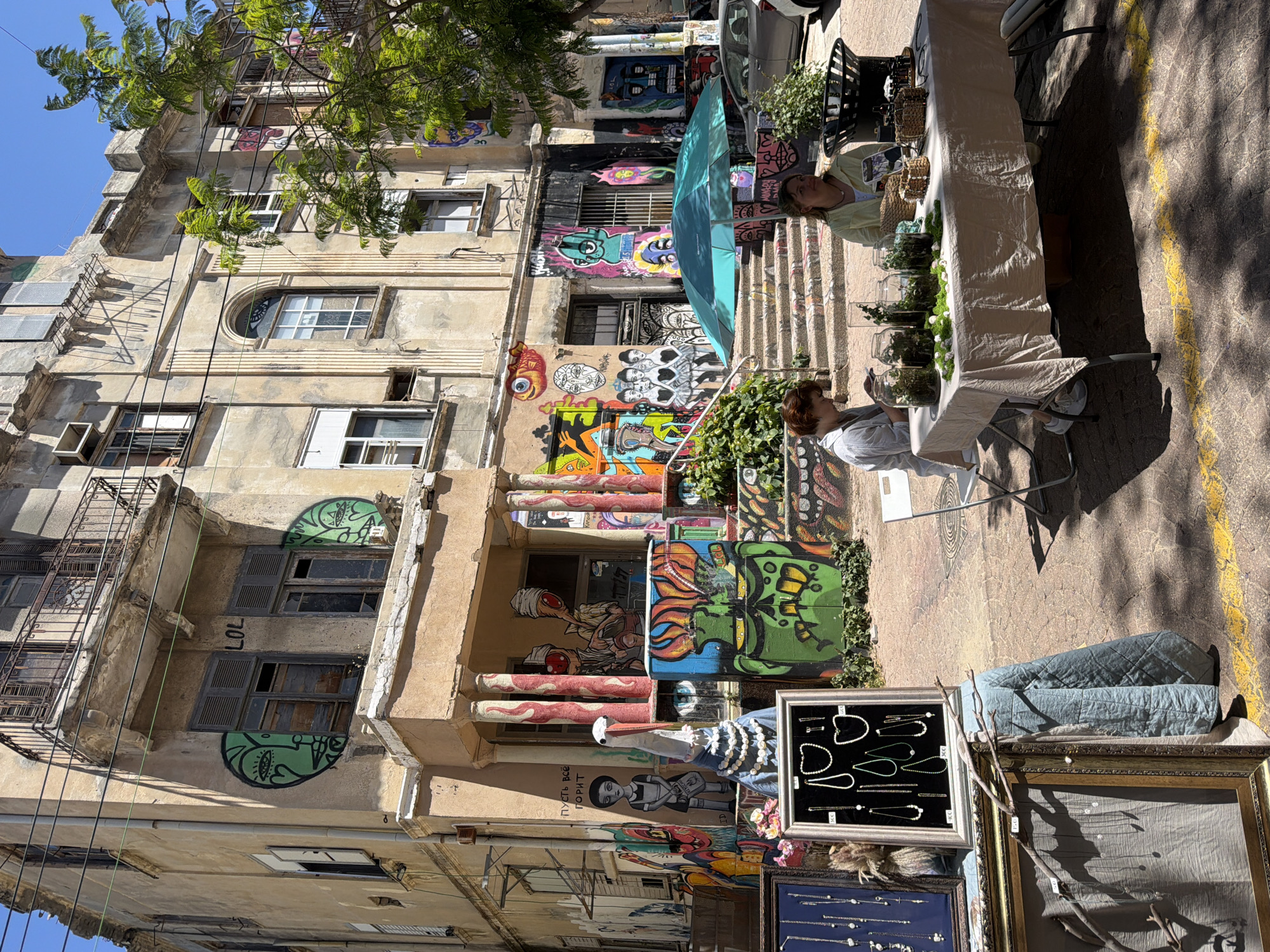 The image shows a street scene in Tel Aviv, Israel, likely in the Florentin neighborhood, known for its street art. An old, multi-story building with graffiti-covered walls forms the backdrop. The building appears aged with various architectural details. The street is lined with vendors. A table covered with a white tablecloth is partially visible on the right side of the frame, appearing to display terrariums with greenery inside. Two people are near the table; one is seated and looking at a phone, while the other stands behind the table. Below the building, on the street, there are displays of jewelry. The ground has a marked yellow line. A tree branches out at the top of the frame.