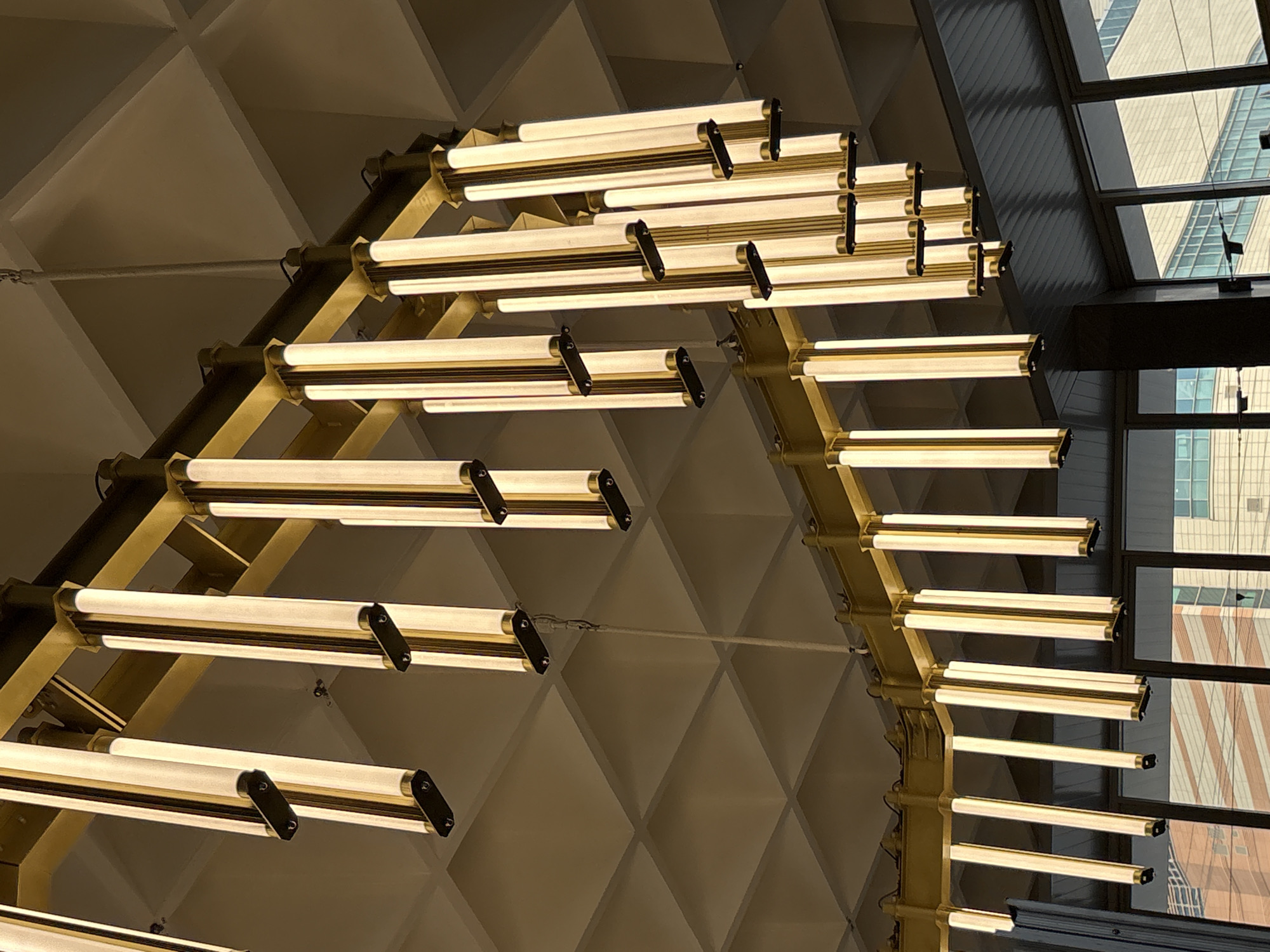 The image shows a grand chandelier with multiple vertical glass tubes illuminated with warm light, suspended from a golden metal frame. The chandelier is mounted on a large, coffered ceiling made of light-colored panels, creating a diamond pattern. Several hanging wires are visible, connecting the chandelier to the ceiling. The lower part of the image reveals a dark, reflective surface, possibly a window or a railing, with a blurry view of buildings and windows in the background. The lighting in the scene is primarily from the chandelier itself, casting a warm glow. There are no people visible in the image. The architectural style and the elaborate chandelier suggest a formal or public building, possibly a theater, hotel lobby, or a museum. However, the exact location in Saint Petersburg, Russia, cannot be determined from the visual information provided.