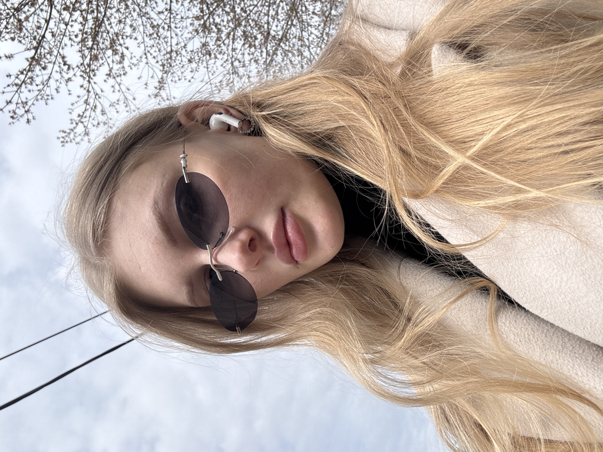 A woman with blond hair is wearing sunglasses and an earbud. The background includes bare tree branches and the sky. Power lines are visible. The picture was taken in Perm, Russia.