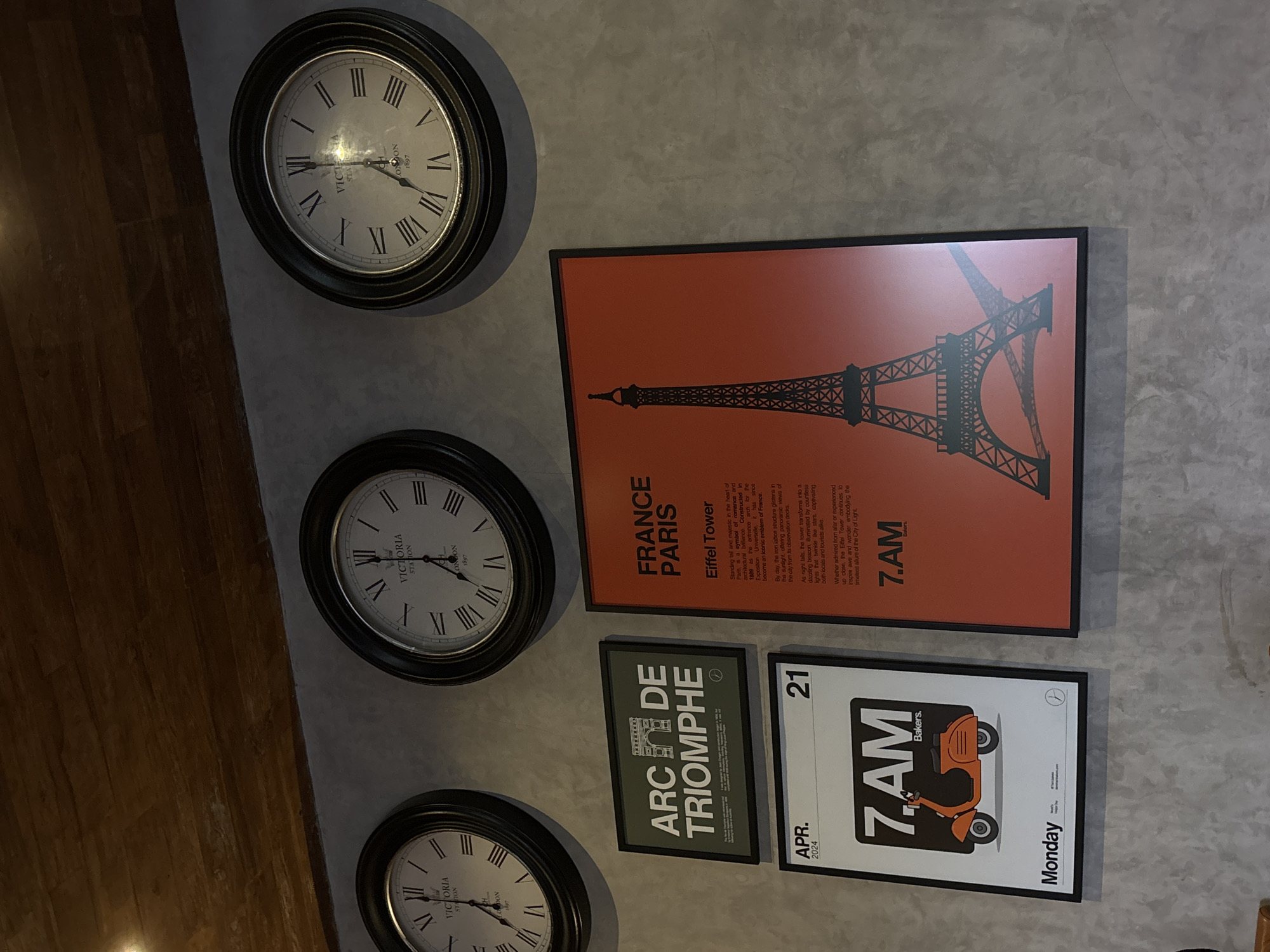 This image captures a wall adorned with travel-themed decorations in Mengwi, Indonesia. The wall features a textured, light gray concrete finish, with a strip of dark wooden paneling visible on the left side, suggesting an adjacent floor or wall.
Three circular clocks with Roman numerals are mounted vertically along the left side of the wall. Each clock has a black frame, a white face, and black hands. The text "VICTORIA STATION LONDON 1897" is visible on the faces of all three clocks. The top clock shows approximately 1:50, the middle clock shows approximately 3:20, and the bottom clock shows approximately 7:35.
To the right of the clocks, three framed posters are arranged vertically. The largest, top poster has a vibrant orange background with a black silhouette of the Eiffel Tower. Text on this poster reads "FRANCE PARIS" and "Eiffel Tower," followed by a detailed description of the monument's history, architecture, and appearance during day and night. The text also includes "7.AM" in the bottom right corner.
Below the Eiffel Tower poster is a smaller, rectangular poster with a dark green background and white text that says "ARC DE TRIOMPHE," accompanied by a simplified graphic of the landmark.
The bottommost poster is primarily white with an orange retro-style scooter depicted centrally. Visible text on this poster includes "APR. 2024," "21," "7.AM Bakers.," and "Monday." The overall scene creates an atmosphere of travel and time. The lighting suggests an indoor setting, possibly with artificial light, given the slight glare on the clock faces.
