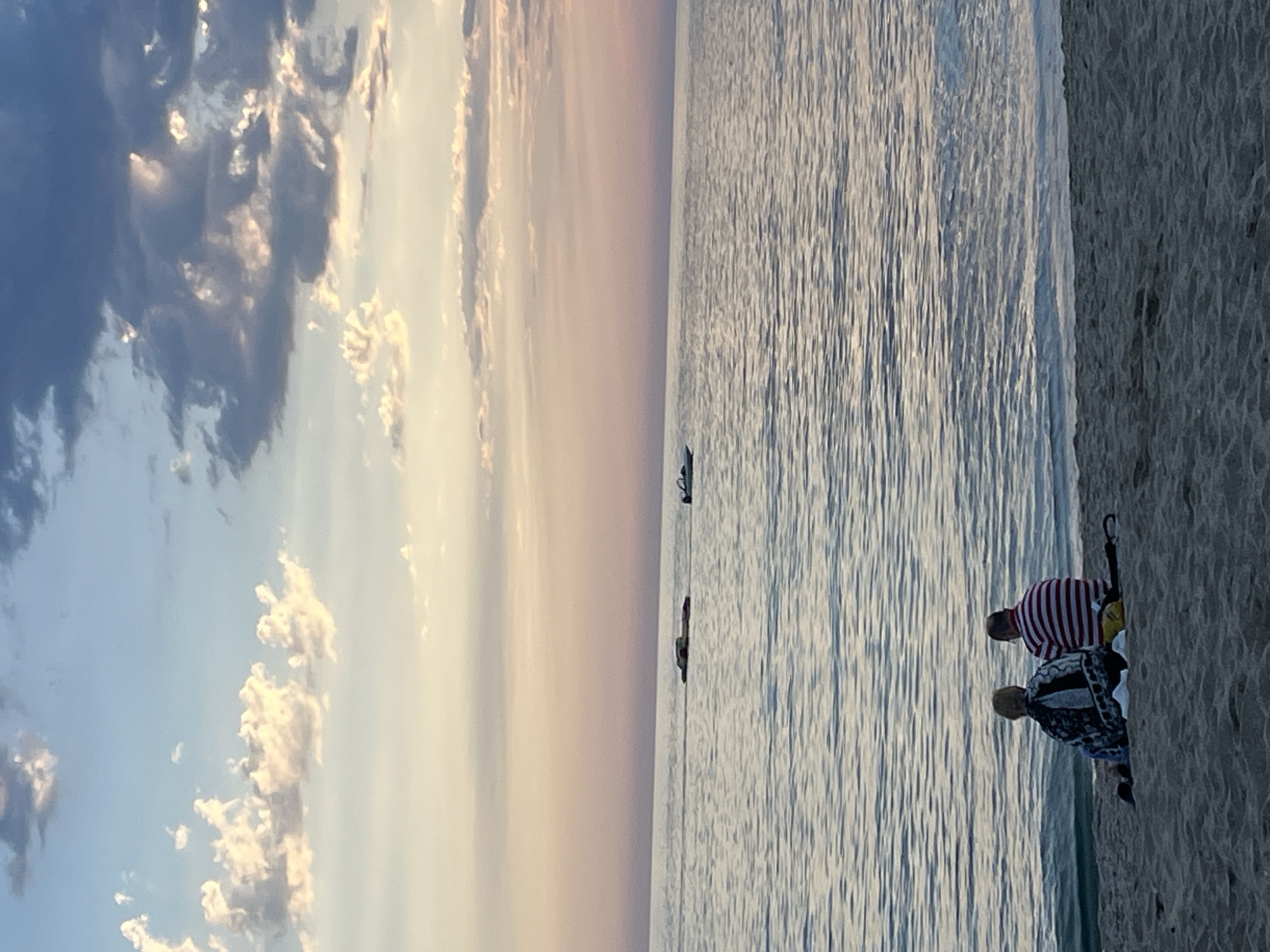 The image shows a beach scene in Alanya, Turkey at dusk. The sky is divided into thirds, with dark clouds on the left, a soft sunset hue in the center, and the sea on the right. There are two small boats on the water. On the beach, two people stand near the waterline; one is wearing a red and white striped shirt, the other a patterned jacket. An open umbrella lies on the sand next to them.
