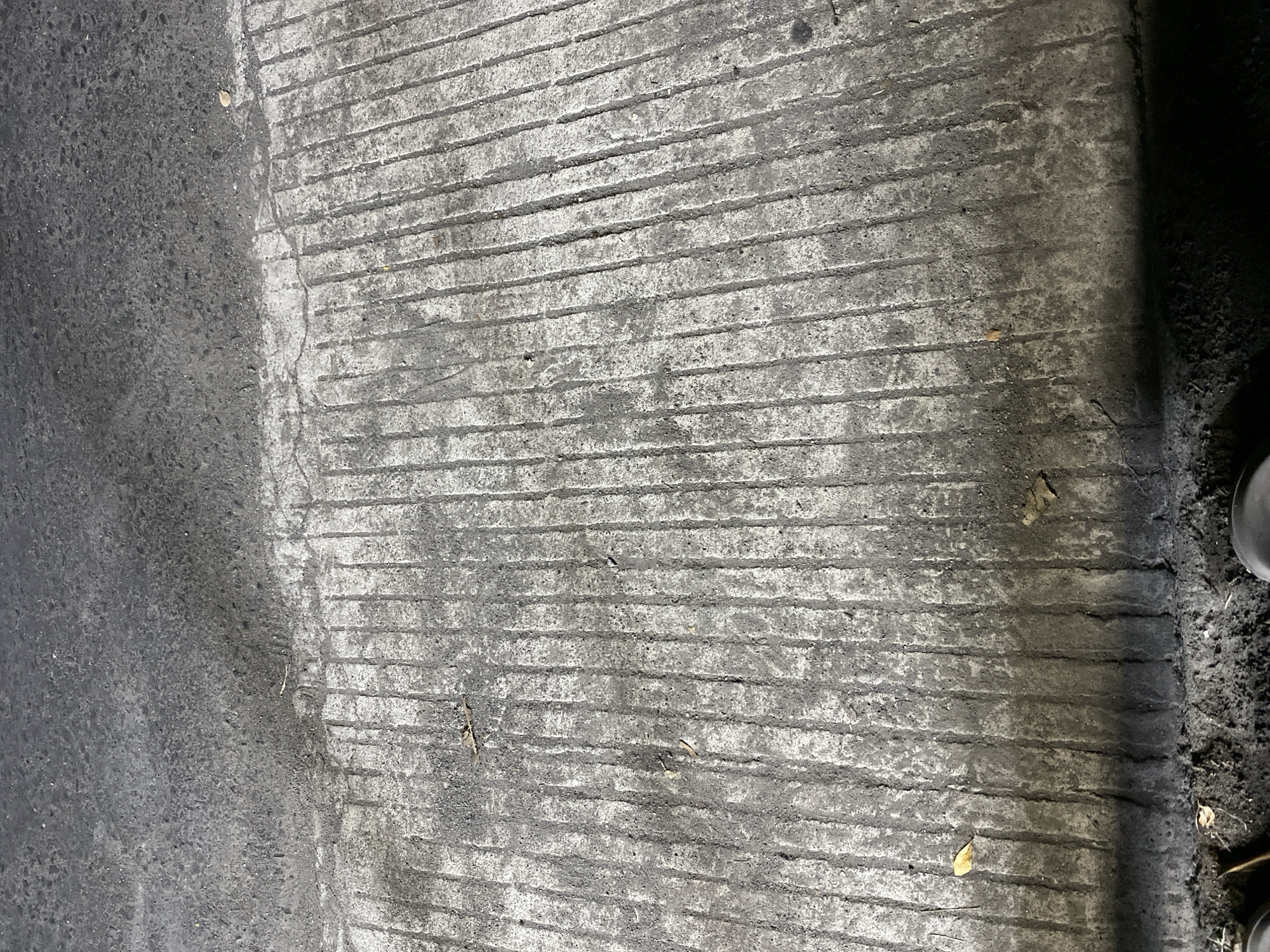 A high-angle close-up shot captures a section of a textured concrete surface, possibly a sidewalk or pathway, in Kasihan, Indonesia. The concrete is distinguished by deep, parallel grooves running vertically down the frame, creating a ribbed appearance. The surface is stained and weathered, with areas of lighter grey concrete interspersed with darker, grimy patches. Scattered leaves and debris, including at least two brown, dry leaves, are visible on the concrete. The top of the image shows a transition to a smoother, darker asphalt surface, suggesting a change in material or a different part of the roadway. The overall impression is one of an outdoor, urban environment, with the focus being on the tactile and visual details of the ground surface.