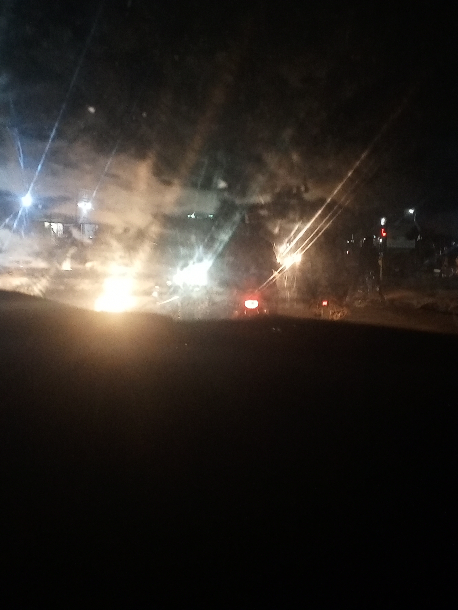 The image depicts a nighttime scene with obscured visibility. Bright light sources, likely headlights and streetlights, create significant glare. A building is visible in the upper left, and traffic signals are present on the right. A vehicle with red taillights is also visible. The lower portion of the image is mostly dark, obscuring the foreground details. The location is in the city of Unknown, Unknown.