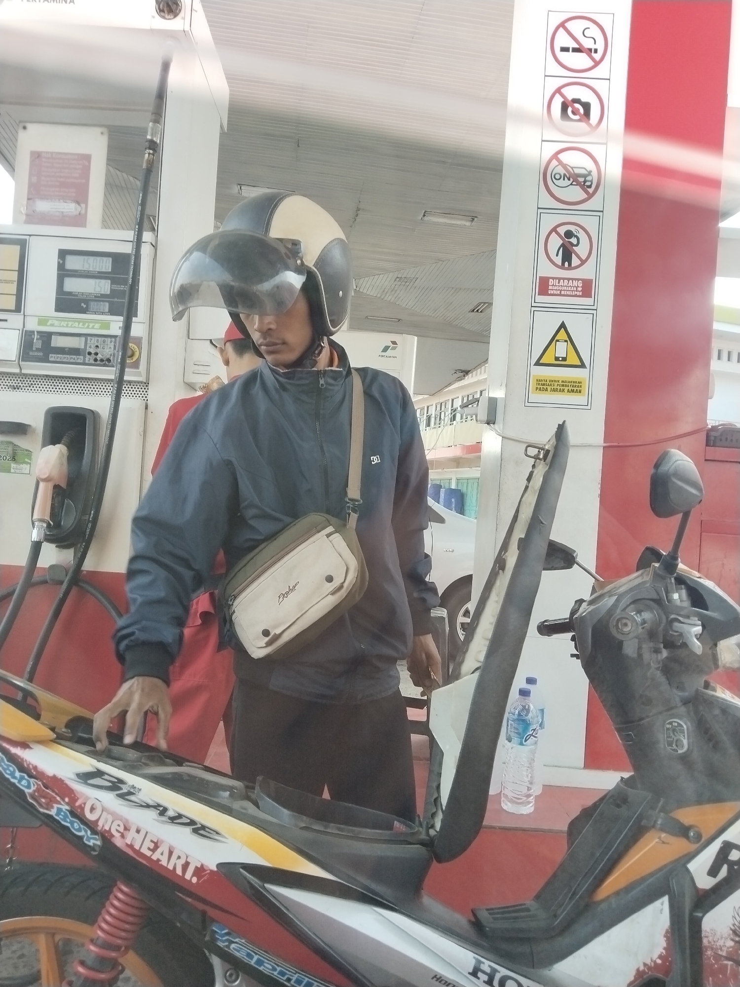 In Adiwerna, Indonesia, at a Pertamina gas station, a man wearing a blue jacket, black pants, and a helmet with a clear visor stands next to his motorcycle. He has a cross-body bag with beige and olive green compartments. The man is reaching towards the motorcycle's seat, which is partially open. A gas pump nozzle is visible near him, and a bottle of water rests on the ground beside the motorcycle. In the background, another person wearing a red uniform is visible, and signage with prohibition symbols and text in Indonesian is displayed on a pillar. The environment appears to be an outdoor, covered area, likely a gas station canopy.