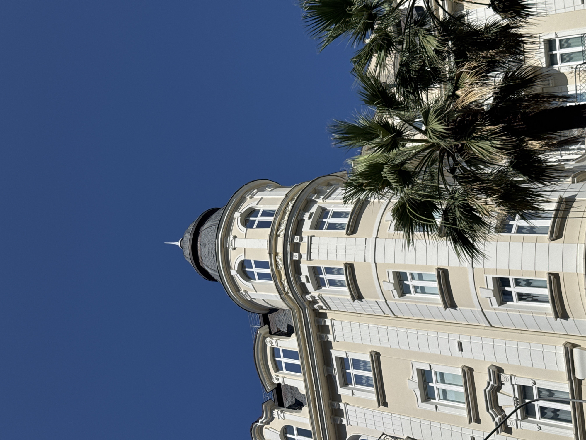 The image shows a low-angle view of an ornate cream-colored building with a dark, conical roof topped with a finial, set against a clear blue sky. The building features multiple windows, some with arched tops and decorative white trim. Palm trees with lush green fronds are partially obscuring the lower section of the building. This architectural style and the presence of palm trees are characteristic of Sochi, Russia, a Black Sea resort city known for its subtropical climate and distinctive architecture.