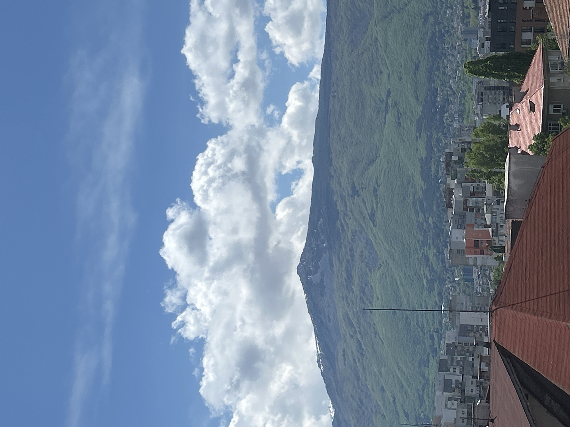 The image shows a mountain partially obscured by clouds, with a portion of the city of Sofia, Bulgaria, visible at the base of the mountain. The sky is blue with fluffy white clouds. Buildings with orange-tiled roofs and other residential and commercial structures are visible. A dark, thin, vertical structure extends upwards from the rooftop. The mountain is green, indicating vegetation, with some areas appearing rocky or bare.