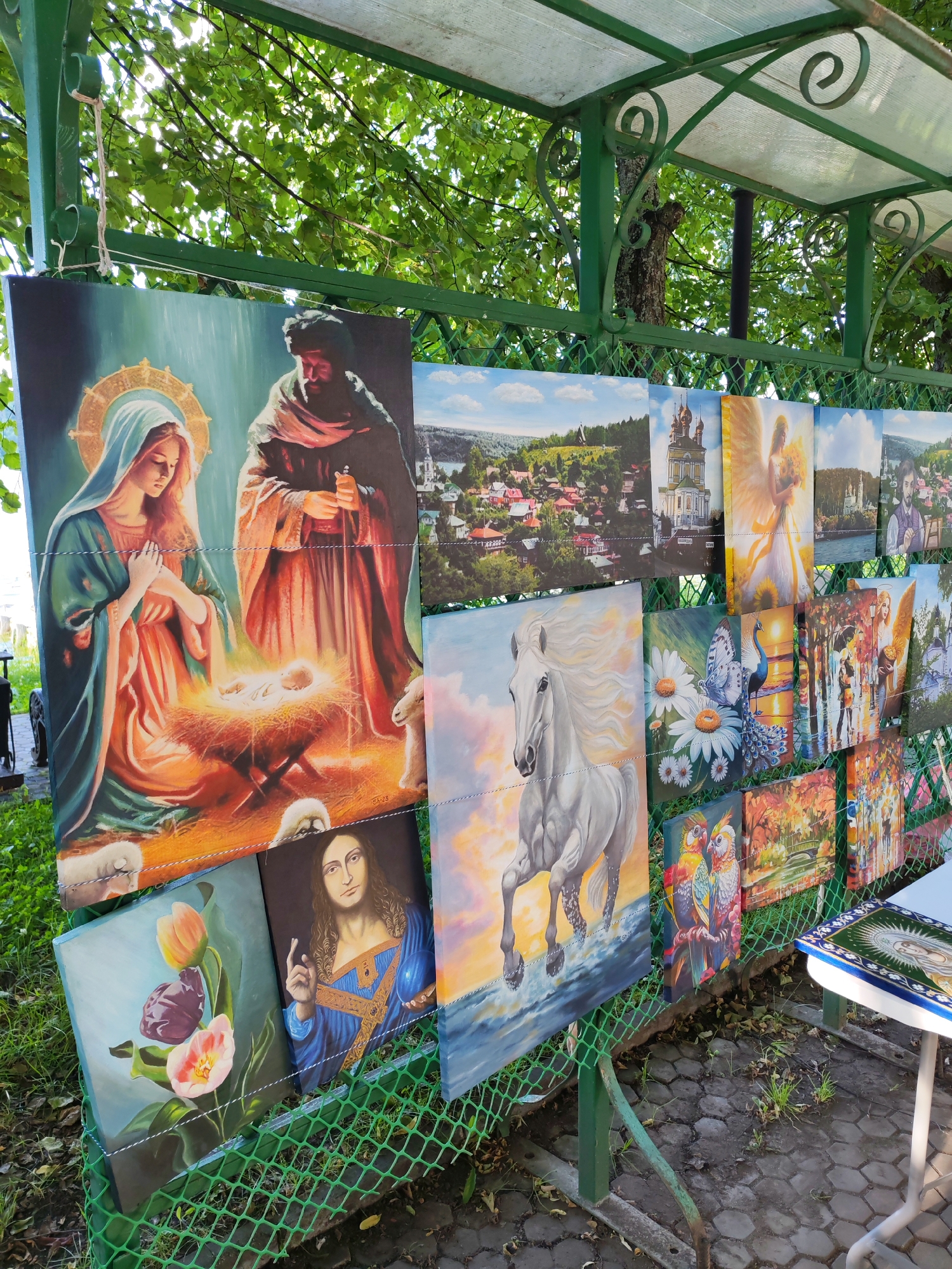 This media file displays a variety of paintings exhibited outdoors, likely at a market or art fair. The paintings are hung on a green metal fence under a covered structure, suggesting an open-air venue. The background includes lush green trees and foliage, indicating a daytime setting, possibly with dappled sunlight filtering through. The ground appears to be paved with hexagonal stones.
The subjects of the paintings are diverse, featuring religious scenes, animals, landscapes, and figures. Prominently displayed is a large painting of the Nativity scene, with Mary, Joseph, and the infant Jesus in a manger, surrounded by sheep. Other paintings include a white horse in motion, a detailed depiction of parrots, a butterfly and peacock with daisies, a serene landscape of a village with a church, an angel holding a bouquet of sunflowers, and a portrait of Jesus Christ. Several other paintings are visible, showing further landscapes and figures.
A small table to the right is partially visible, with a book or album on it featuring a religious image. The overall impression is one of an artist or vendor displaying their work for sale in a picturesque, possibly historic, location. The style of the paintings varies, but many appear to be rendered in a realistic or semi-realistic manner, with vibrant colors and attention to detail. The time of day appears to be daytime, given the brightness and shadows, and the weather seems pleasant, conducive to an outdoor exhibition.
The signature "Shiz" is visible on the Nativity painting.