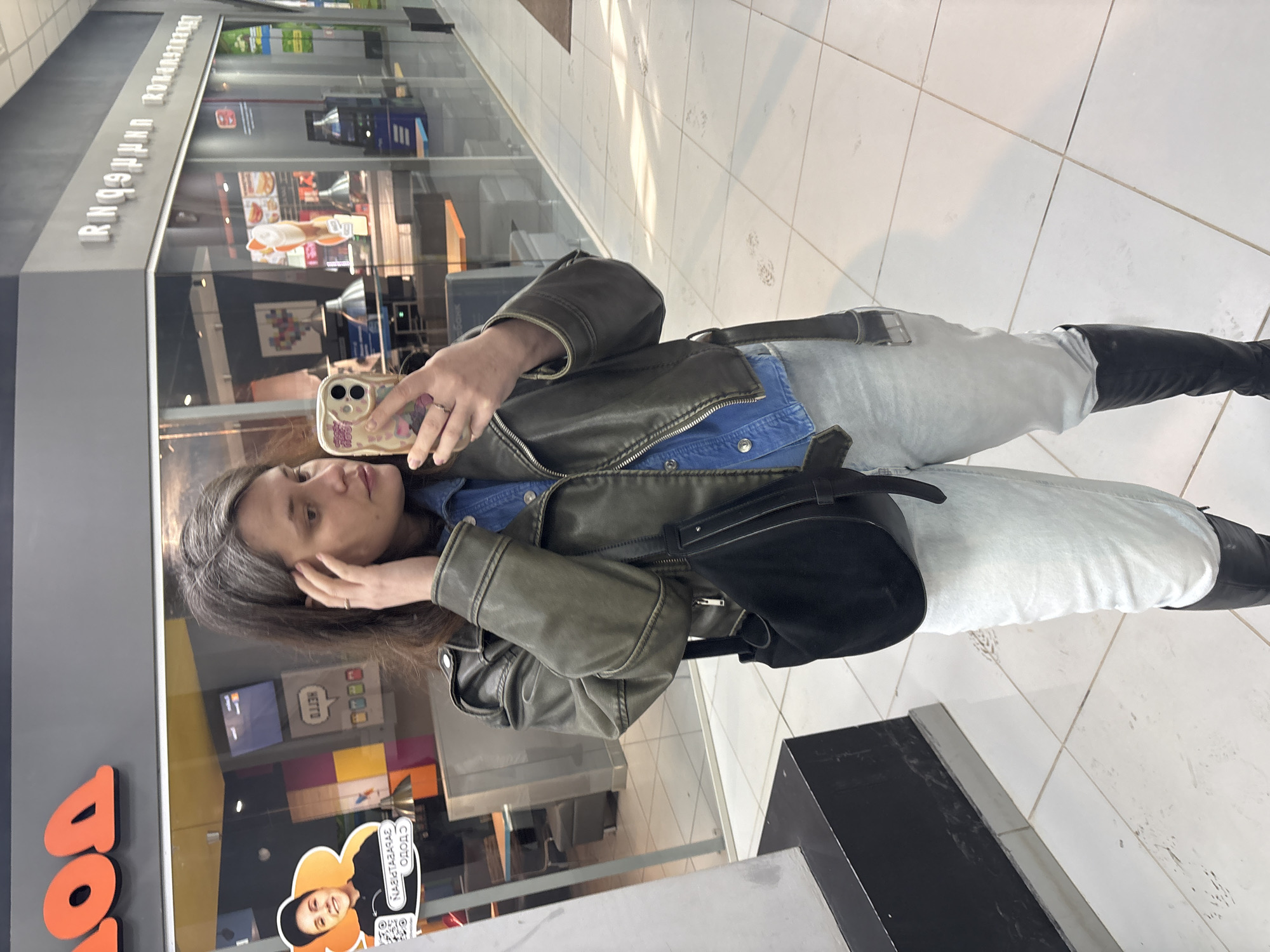 An adult female is depicted taking a mirror selfie inside a commercial establishment, likely a shopping mall or food court, located in Izhevsk, Russia. The individual has long, dark brown hair with noticeable graying and is wearing a dark olive-green zippered motorcycle-style jacket over a light blue denim shirt, light-wash blue jeans, and tall black boots. A black hobo-style shoulder bag with a wide strap is slung over her right shoulder. She holds a light-colored smartphone with a patterned case in her right hand, positioned to capture her reflection, while her left hand touches her temple. A ring is visible on the ring finger of her right hand.
The background features multiple glass storefronts reflecting the interior. To the immediate left, partial orange Cyrillic letters "ДО" are visible, corresponding to a "Додо Пицца" sign. Adjacent to this, a promotional standee displays an image of a person in a black cap with Cyrillic text "НАВИТАЗАРБА" and "ОДОДО" and a QR code. To the right, inverted Cyrillic lettering "ЯПОНИЯ-ГОРОД" (Japan City) is visible above another food establishment. The reflected shop interiors reveal tables, menu screens, and food advertisements. The floor is composed of large, light-colored square tiles, exhibiting some scuff marks and dirt. The scene is illuminated by overhead artificial lighting.