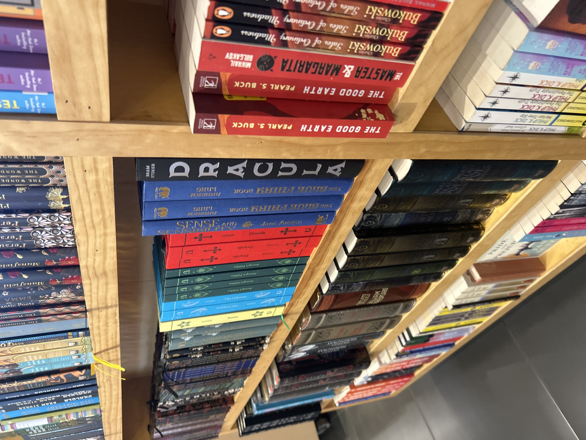 This is a close-up shot of a bookshelf, likely within a bookstore. The shelves are made of light-colored wood and are packed with books of varying sizes, colors, and titles. The books are arranged in rows, with spines facing outward. Notable titles visible include "The Good Earth" by Pearl S. Buck, "The Master and Margarita" by Mikhail Bulgakov, various works by Charles Bukowski ("Tales of Ordinary Madness"), "Dracula" by Bram Stoker, "Sense and Sensibility" by Jane Austen, and "The Blue Fairy Book" by Andrew Lang. There are also numerous copies of works by Philip K. Dick. The lighting is bright and even, allowing for clear visibility of the book titles and covers. A yellow ribbon is used as a bookmark in a book on the left side of the image. The books are stacked tightly on the shelf, suggesting a well-stocked or possibly even overcrowded bookstore. The location is implied to be in the city of Unknown, Unknown.