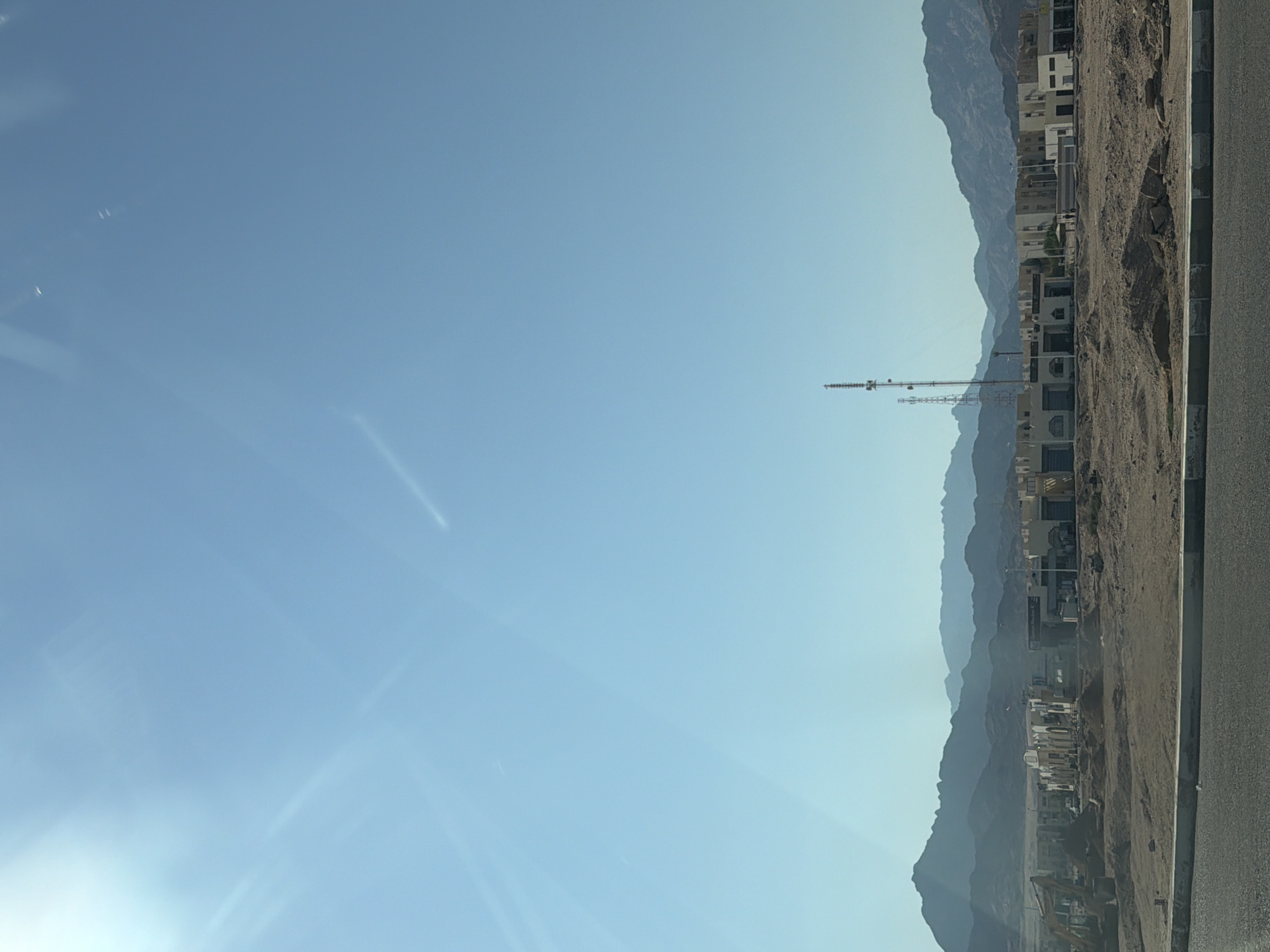 The image shows a desert landscape with mountains in the background. In the foreground, there is a road and a construction site. There are several buildings under construction, and some appear to be shops or small businesses. Two telecommunication towers are visible in the middle ground. The sky is clear and blue, with some lens flare from the sun. The overall scene depicts a developing area in a desert environment.