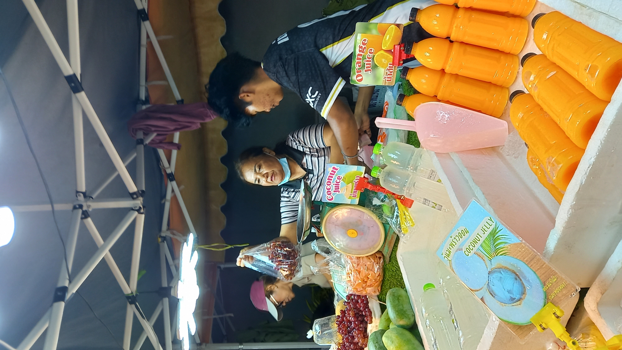 This image depicts a street food stall in Koh Phangan, Thailand, specializing in juices and fruits. Two vendors, a woman and a young man, are visible behind a display of beverages and fresh produce. The woman, wearing a striped shirt and a light blue mask, is weighing what appears to be dried fruit in a plastic bag on a green scale. In front of them, a cooler is filled with bottles of orange juice and what looks like coconut jelly, along with some fresh mangoes and grapes. Signage indicates "Coconut Juice" and "Orange Juice" in English and Thai. The stall is set up under a white tent structure, illuminated by artificial lights, suggesting it is operating in the evening or at night. The background shows a glimpse of a building and foliage, consistent with a tropical outdoor market setting.