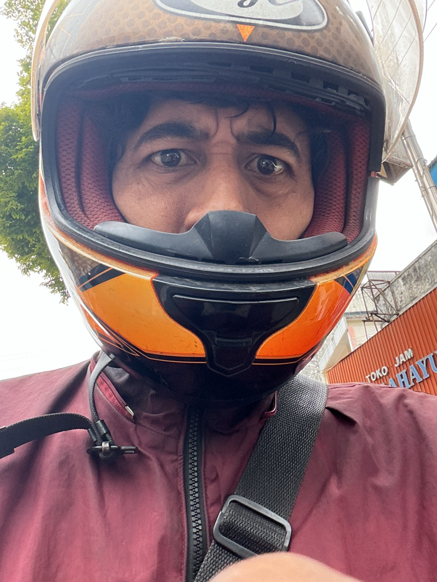 A person wearing an orange and black motorcycle helmet and a maroon jacket is captured in a close-up, front-facing selfie. The helmet's interior lining is visible as red, quilted fabric. A black strap from a bag crosses the person's chest. In the background, a building with an orange corrugated metal facade is partially visible, with lettering that reads "TOKO JAM MAHAYU". Trees and a grey sky are also present in the background. The scene suggests an outdoor urban environment, possibly in Magelang, Indonesia, given the signage.