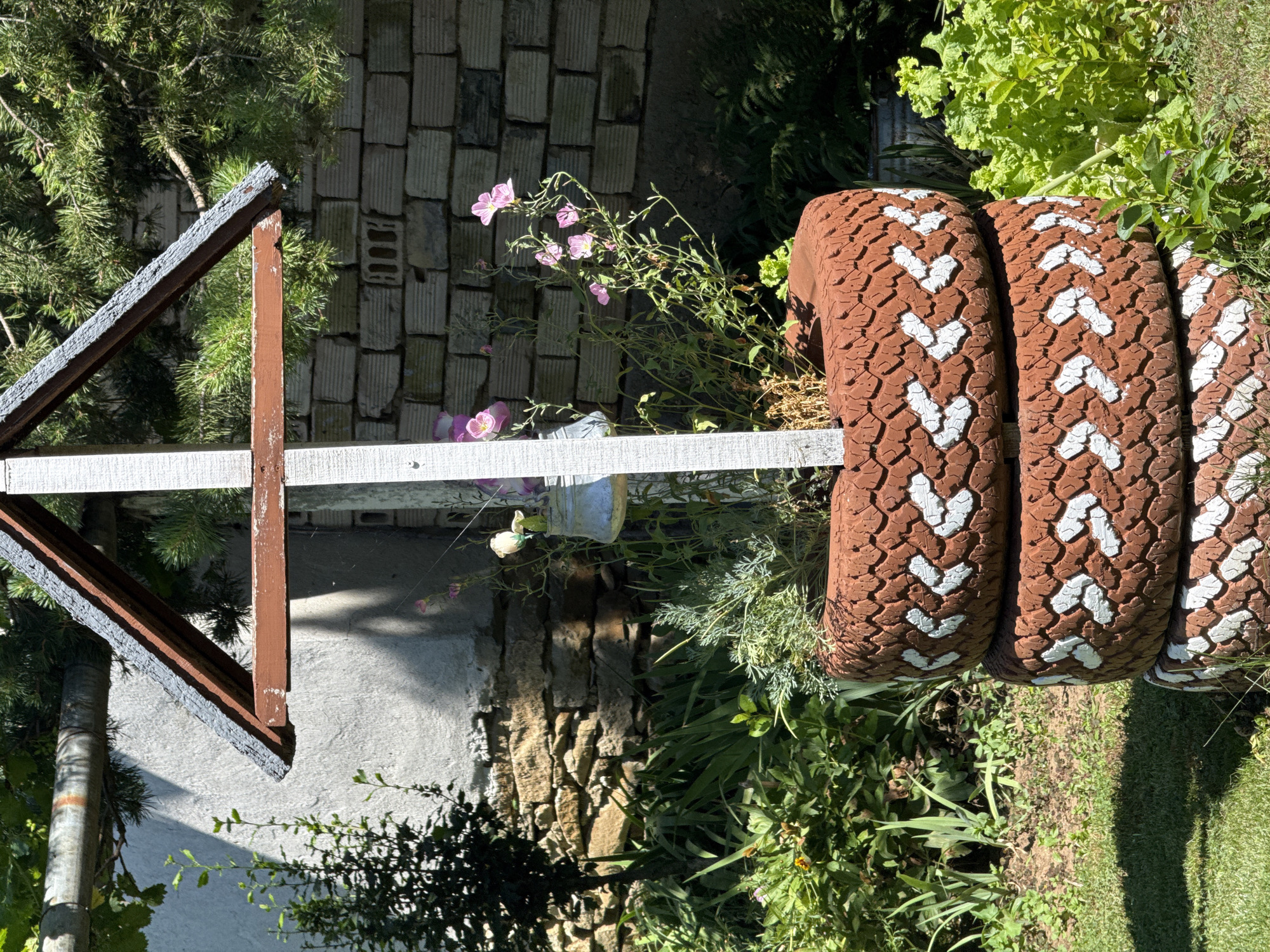 The image shows a decorative garden feature made from three stacked, painted tires. The tires are painted reddish-brown with white chevron patterns around their sides. In the center of the tires, a small wooden structure with a peaked roof has been built. Various plants, including green foliage and some pink flowers, are growing out of the top tire. A small metal watering can hangs from the wooden structure. In the background, a brick wall and a stone wall are visible, along with a large tree. The scene is set in an outdoor garden. The location is Konop, Bulgaria.