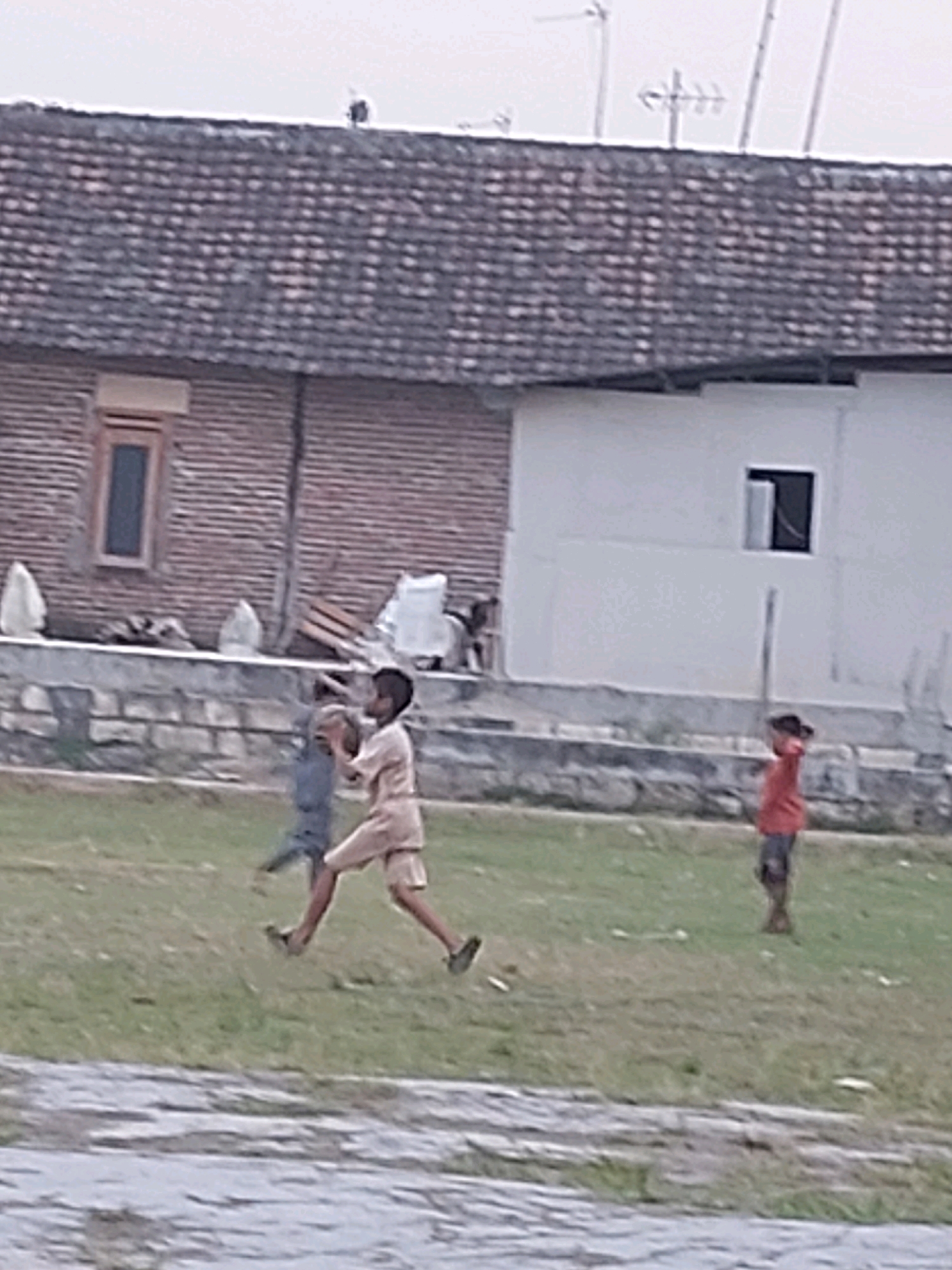 The image shows a slightly blurry, outdoor scene with three young boys in a grassy area in front of buildings. The boy in the center, wearing a light-colored outfit, is running and appears to be holding something in his hands. To his left, another boy in a dark blue outfit is also running. To the right, a boy in a red shirt and dark shorts is standing. In the background, there is a building with a tiled roof and a brick wall, and a white building with a window. There is also a low stone wall separating the grassy area from the buildings. The environment suggests a residential or communal area, possibly within the city of Sarangan, Indonesia, given the architectural style and the presence of people in a casual outdoor setting.