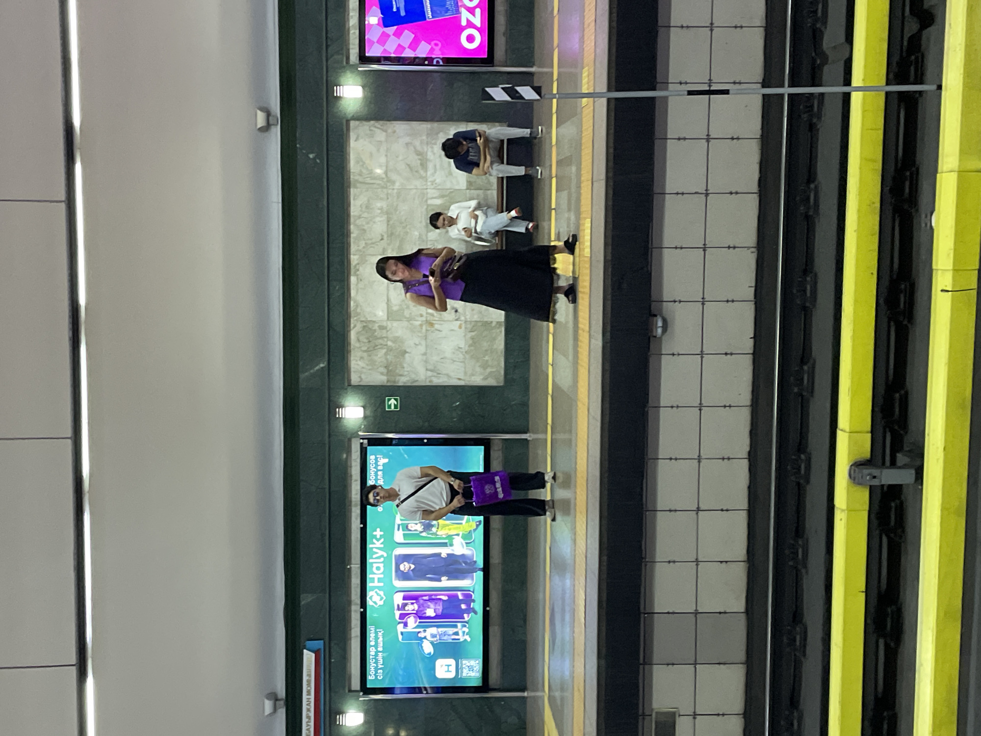 The image shows a subway platform in Almaty, Kazakhstan. In the foreground, there are train tracks with yellow safety markings. The platform has a tiled wall with advertisements displayed on digital screens.
On the platform, several individuals are visible:
* A man wearing a white polo shirt, dark pants, and sunglasses is standing near an advertisement for Halyk+. He is holding a purple shopping bag.
* A woman in a purple sleeveless top and a long black skirt is standing and looking at her phone.
* Two other individuals are seated on a bench in the background. One person is wearing a white top and jeans, and the other is wearing a grey t-shirt and pants.
* Another advertisement for "OZON" is visible on a pink screen on the right side of the image.
The environment appears to be an indoor subway station, indicated by the overhead lighting and the enclosed architecture. The lighting is a mix of fluorescent lights and the glow from the digital advertisements. The overall scene depicts a typical public transportation waiting area.