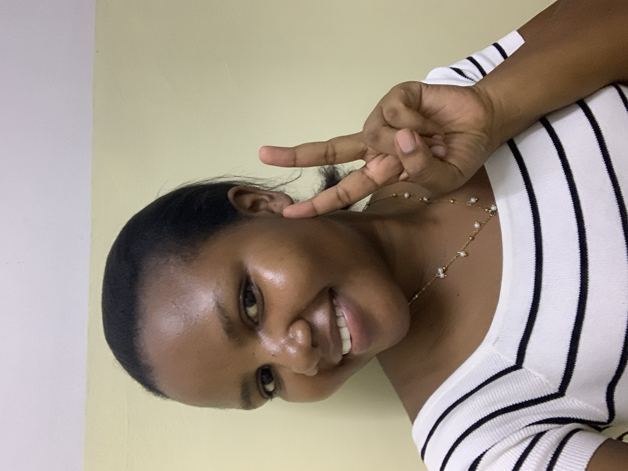 A young woman is posing for a portrait indoors. She is wearing a white top with black stripes and a delicate gold necklace with small pearl-like beads. Her dark hair is pulled back, and she is smiling with her teeth showing, making a peace sign with her right hand near her right ear. The background is a plain, light yellowish-beige wall. It is impossible to determine if the photo was taken in the city of Dar es-Salaam, Tanzania from the image alone.