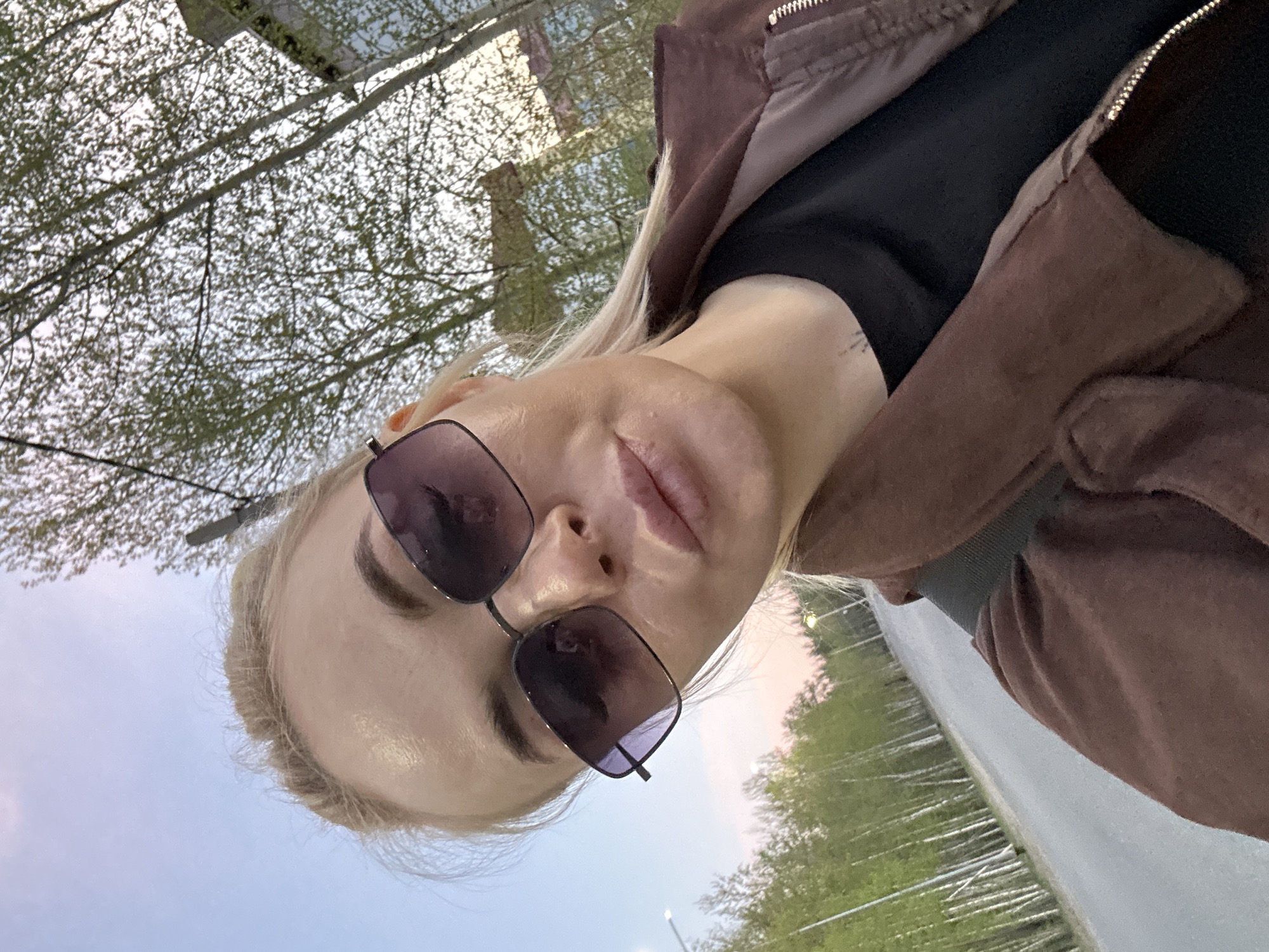 A woman is wearing sunglasses, a dark top and a brown jacket. Trees and a paved surface are visible in the background. The photo was taken in Vysoky, Russia.