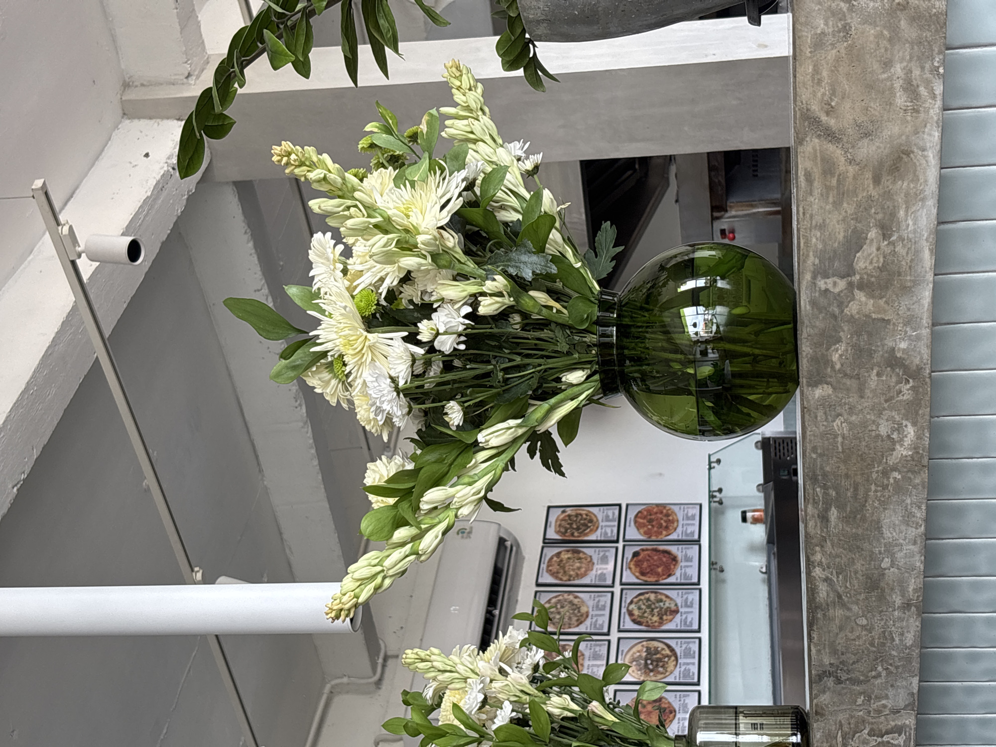A close-up, eye-level shot captures a large bouquet of white flowers, primarily chrysanthemums and what appear to be tuberose, arranged in a round, dark green glass vase. The vase sits on a textured, light gray concrete counter. Behind the flowers, a wall displays a grid of framed pictures, each showing a different pizza. Above this, a white air conditioning unit is visible, and further up, a white cylindrical object hangs from the ceiling. The background suggests an indoor setting, possibly a restaurant or cafe, with industrial-style ceilings and exposed beams. The scene is lit with artificial light, creating soft shadows. No people are visible in the frame. The image has a shallow depth of field, with the flowers and vase in sharp focus and the background slightly blurred.