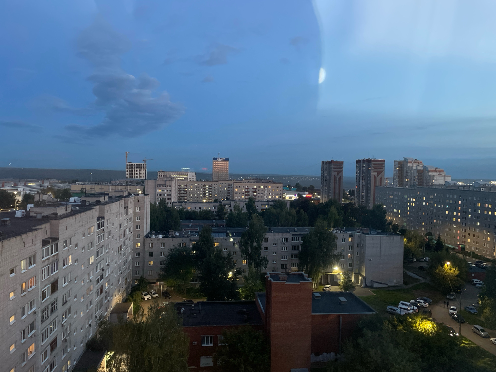 This is an aerial view of a residential area in Izhevsk, Russia, at dusk. The scene is dominated by multi-story apartment buildings, characteristic of Soviet-era urban planning. The sky is a deep blue, with some scattered clouds, and a faint reflection of what appears to be a moon or a light source is visible on the right side of the image.
In the foreground, a long, pale grey apartment building with many windows is prominent. The windows show a mix of illuminated and dark panes, indicating that some residents are home. Adjacent to this building, other apartment blocks of similar design are visible, interspersed with trees. Some of the buildings are illuminated by streetlights, casting a yellowish glow on the surrounding areas.
In the middle ground, more apartment buildings of varying heights and architectural styles are present. Some are older, grey concrete structures, while others are taller, more modern buildings with balconies and colorful facades. A construction crane is visible near a tall building on the left side of the image, suggesting ongoing development. Trees and vegetation are scattered throughout the urban landscape, softening the concrete structures.
In the background, the city extends further, with more buildings and lights visible in the distance. A dark, forested area or a hill forms a horizontal line on the horizon. Several cars are parked along streets and in courtyards within the residential complex. A few individuals can be seen walking on a street, but they are too small to be identified. The overall atmosphere is one of a typical evening in a large Russian city.