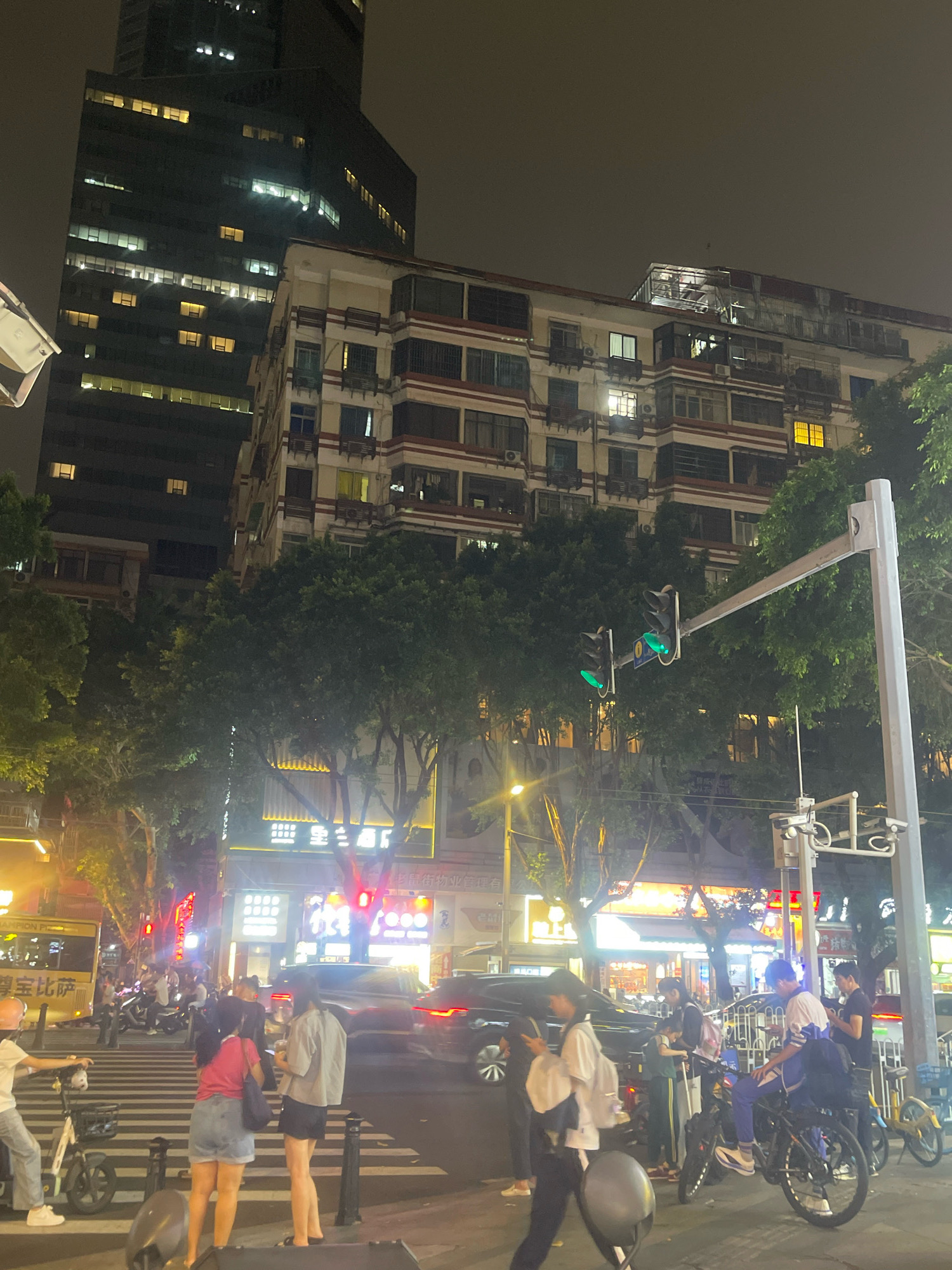 At nighttime in Yuexiu District, China, an intersection bustles with activity. A tall, modern skyscraper looms in the background, its windows illuminated, contrasting with a more aged, multi-story building featuring a red and white striped facade. Trees line the street, their foliage partially obscuring the buildings. Traffic lights, showing green, hang over the road. A pedestrian crossing is visible, with several people on foot and on bicycles. A yellow bus is parked on the left, its side displaying Chinese characters and a slogan. Various vehicles, including cars and motorcycles, are present, indicating vehicular traffic. The scene is illuminated by streetlights and the glow from buildings and signs.