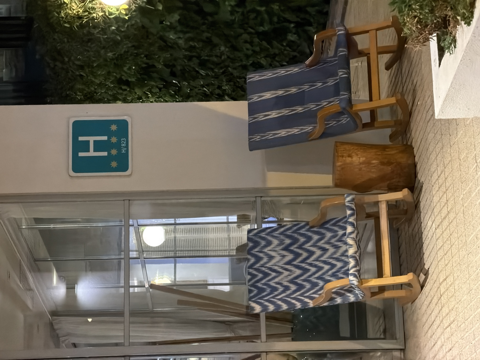 The image captures an outdoor scene in Palmanova, Spain, at dusk. Two wooden rocking chairs, one with a blue and white zig-zag patterned cushion and the other with a blue and white striped cushion, are placed on a tiled patio. A rustic wooden log serves as a small table between the chairs. Behind the chairs, a large window with sheer curtains reflects light. A teal sign with a white "H" and three stars, indicating a hotel, is mounted on a white wall to the right of the window. Lush green foliage borders the right side of the frame, with a warm-toned outdoor light fixture visible. The lighting suggests it is evening or nighttime.