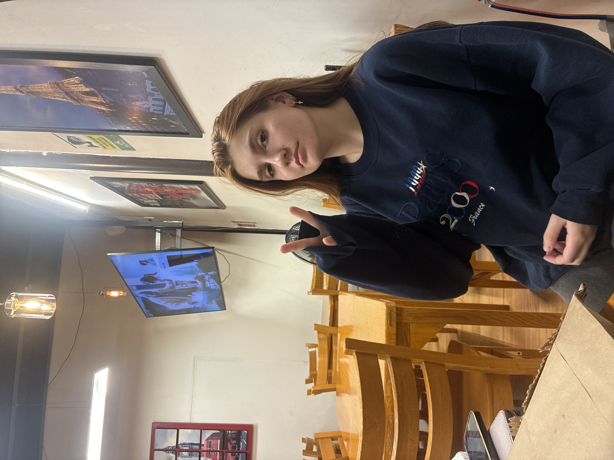 A young woman with auburn hair is sitting at a wooden table inside a restaurant. She is wearing a navy blue sweatshirt with the embroidered text "Paris 2024 France" and making a peace sign with her left hand. The restaurant features wooden chairs, tables, and framed pictures of landmarks on the walls. A television is mounted near the ceiling, and hanging light fixtures are visible. A paper bag and a cell phone are on the table. The specific location of Huixquilucan de Degollado, Mexico is not discernable from the image.