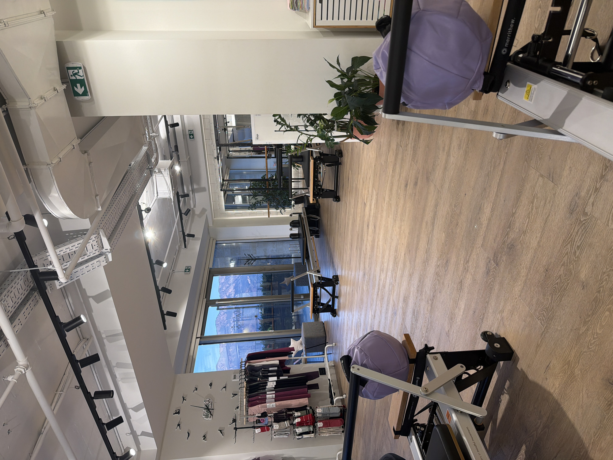 The image depicts the interior of a fitness studio, likely a Pilates studio, located in Boreti, Montenegro. The studio features polished wooden floors and large windows offering a view of mountains and water.
In the foreground, two Pilates reformers are visible. The one on the left is partially obscured, but its metallic frame, black seat, and a large, light purple weighted ball are discernible. The reformer on the right is more fully visible, showcasing its metallic structure, a wooden footrest, a black padded carriage, and a portion of the same light purple weighted ball. A caution sticker is present on the side of this reformer.
The midground reveals more of the studio's layout. A clothing rack with various activewear items is positioned against a wall decorated with a clock and silhouette figures. Beyond this, a seating area with a patterned ottoman is visible. Further into the studio, additional Pilates equipment, including reformers and possibly a Cadillac, are arranged, some with dark metallic frames and black padding. A tall refrigerator or storage unit stands near this equipment, with several potted plants placed in front of it.
The background is dominated by large glass doors and windows looking out onto an exterior space, possibly a patio or balcony, and then the expansive landscape of mountains and water under a sky that suggests either dawn or dusk.
The ceiling is high and industrial, with exposed white pipes, ventilation systems, and black track lighting. Two green illuminated exit signs are visible on the walls. The overall impression is a modern, well-equipped, and well-lit fitness space with an attractive natural backdrop. No people are present in the image.