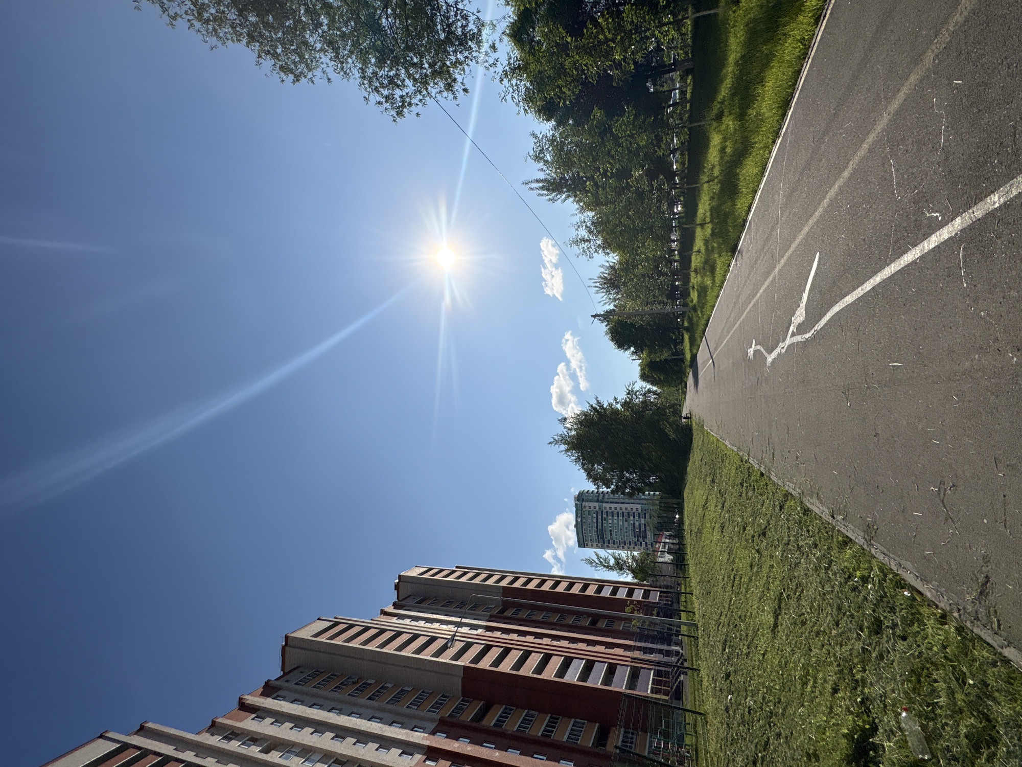 The image captures a bright, sunny day in Izhevsk, Russia, viewed from a low, tilted angle looking upwards towards the sky and surrounding environment. The dominant feature is a brilliant sun, high in a clear blue sky, emanating strong lens flares and rays. A few small, scattered white clouds are visible, and a thin, dark cable or power line stretches across the sky.
On the right side of the frame, a paved path or road, possibly a bicycle lane, runs diagonally, featuring white painted markings including a solid line, a broken line, and a distinct, squiggly white line. Adjacent to this path is a wide strip of vibrant green grass, appearing well-maintained in some areas. A dense line of green trees borders the grass and path, extending into the background.
On the left side, several tall residential buildings are partially visible. The closest building is multi-storied with a distinctive design of alternating light-colored and red-brown sections and numerous windows. Further in the background, another tall, slender modern building stands out against the sky. No people are present in the scene, suggesting a quiet moment in the urban landscape. A small, discarded plastic bottle can be seen on the grass near the path in the lower right. There is no visible text. The overall impression is one of a clear, warm day with strong natural light.
