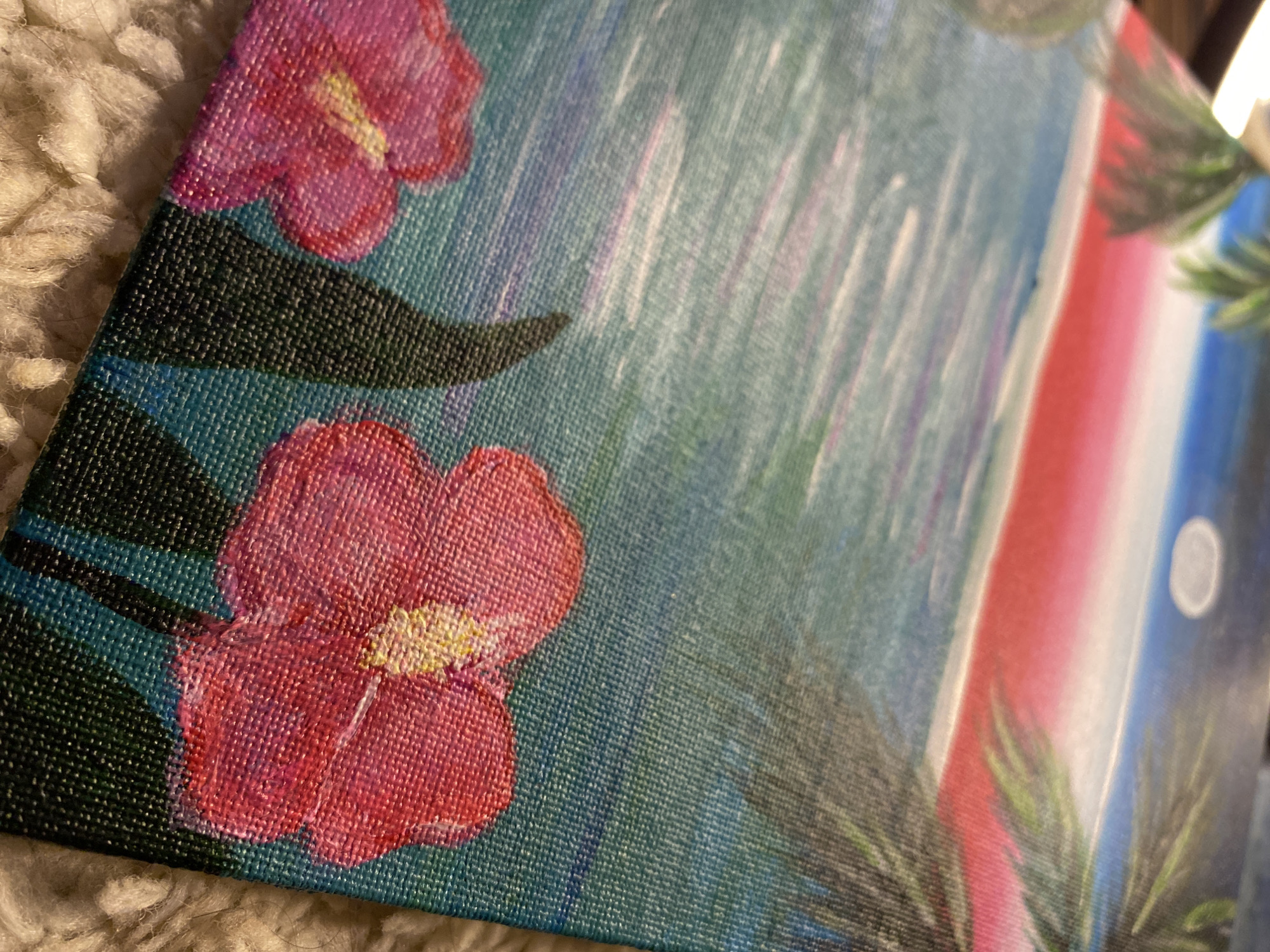 The image shows a close-up of an abstract painting on canvas, likely depicting a tropical scene. Two pink flowers with yellow centers are visible on the left side, surrounded by dark green leaves. The background features a blue and white abstract design, possibly representing a waterfall. Further to the right, a red, white, and blue vertical stripe is present, next to a white circle that is presumed to represent the moon. Some green palm leaves are also depicted on the right side of the canvas. The image does not provide sufficient information to determine if the location is in the city of Chaikovsky, Russia.