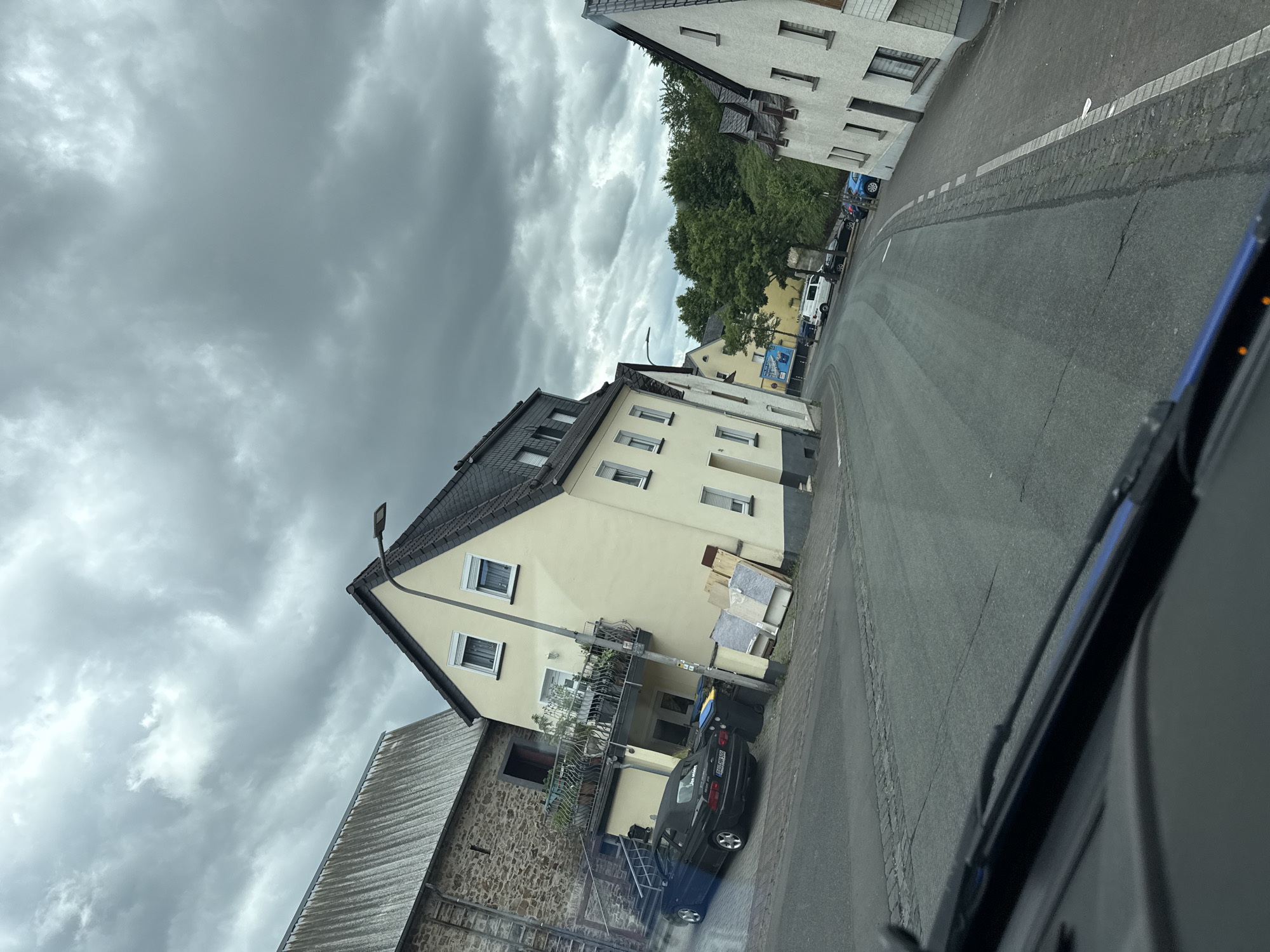 This is a tilted, medium shot of a street in Ulmen, Germany, on a cloudy day. The foreground shows the edge of a car's windshield and wipers. The street is paved with asphalt and has a stone curb. On the left side of the street, there is a light yellow house with a balcony and windows. Next to it is a stone building with a sloped, gray roof. A dark blue convertible is parked in front of the yellow house, and two trash bins are next to it. Further down the street, more houses and trees are visible. On the right side of the street, there is a white house with windows and a slanted roof. The sky is filled with heavy, gray clouds, suggesting an overcast or stormy weather condition.