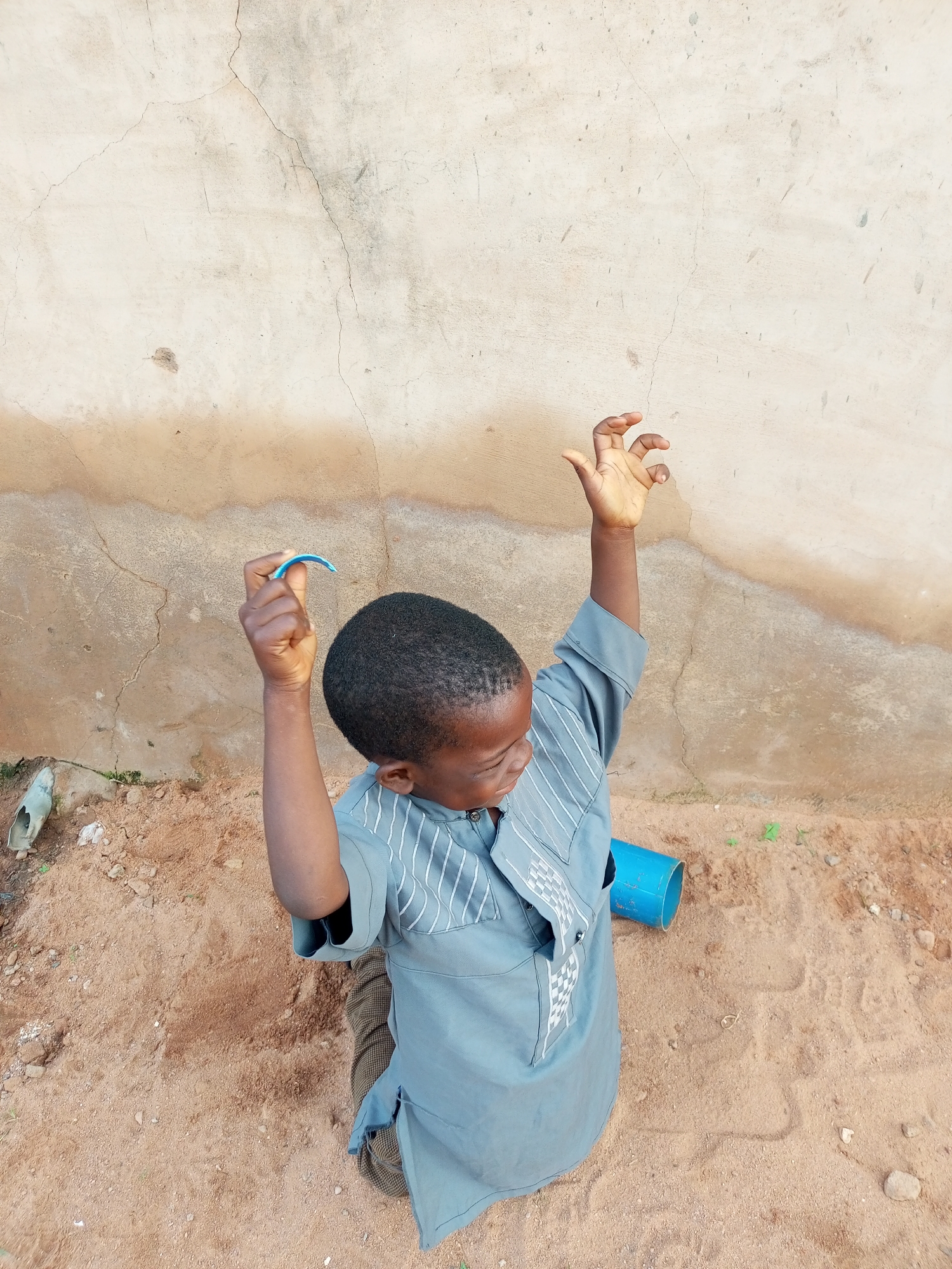 In Tempure, Nigeria, a young Black male is captured from a high angle, kneeling on sandy ground. He is wearing a light blue dashiki with white vertical stripes on the sleeves and chest. His right arm is raised, holding a small blue curved object, possibly a piece of plastic. His left hand is also raised, fingers splayed. The background consists of a textured, light-colored wall with visible cracks and some brown staining near the bottom. Adjacent to the boy on the ground is a blue pipe. The sandy area shows tire tracks and scattered small stones. The lighting suggests it is daytime, casting faint shadows.