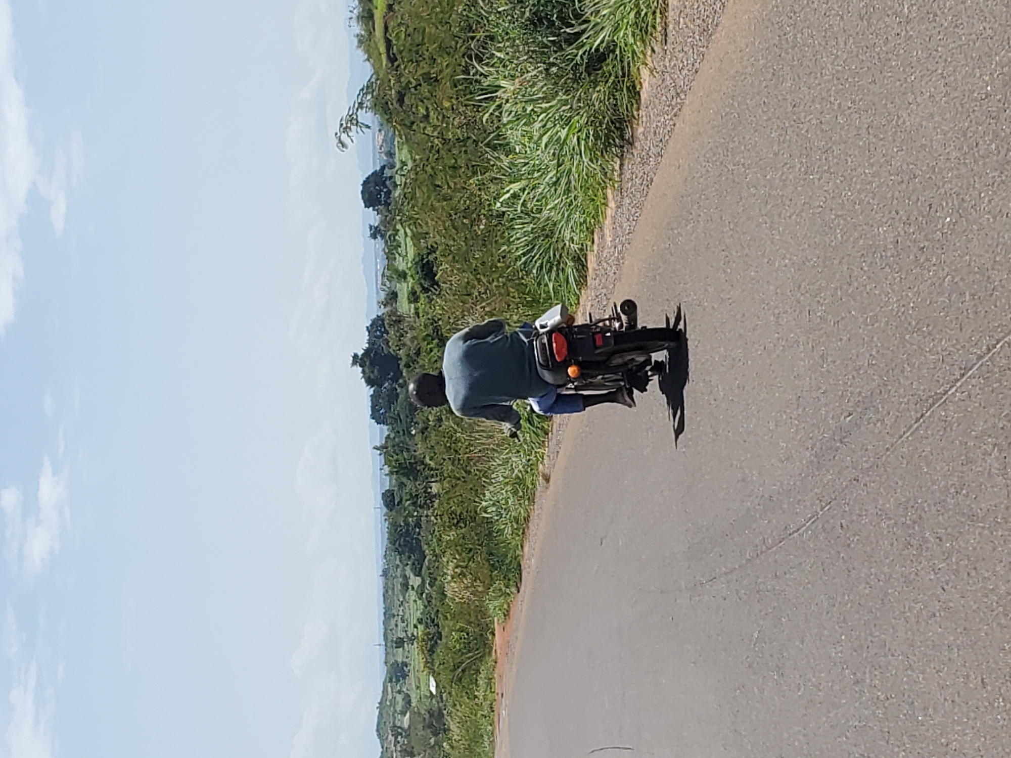 A man wearing a blue shirt and blue pants rides a black motorcycle down a paved road. The motorcycle has a white bag attached to the rear fender. The road is lined with tall green grass on both sides. In the background, there are rolling hills covered in trees and vegetation, with a hazy sky above. The scene appears to be in a rural or semi-rural area.