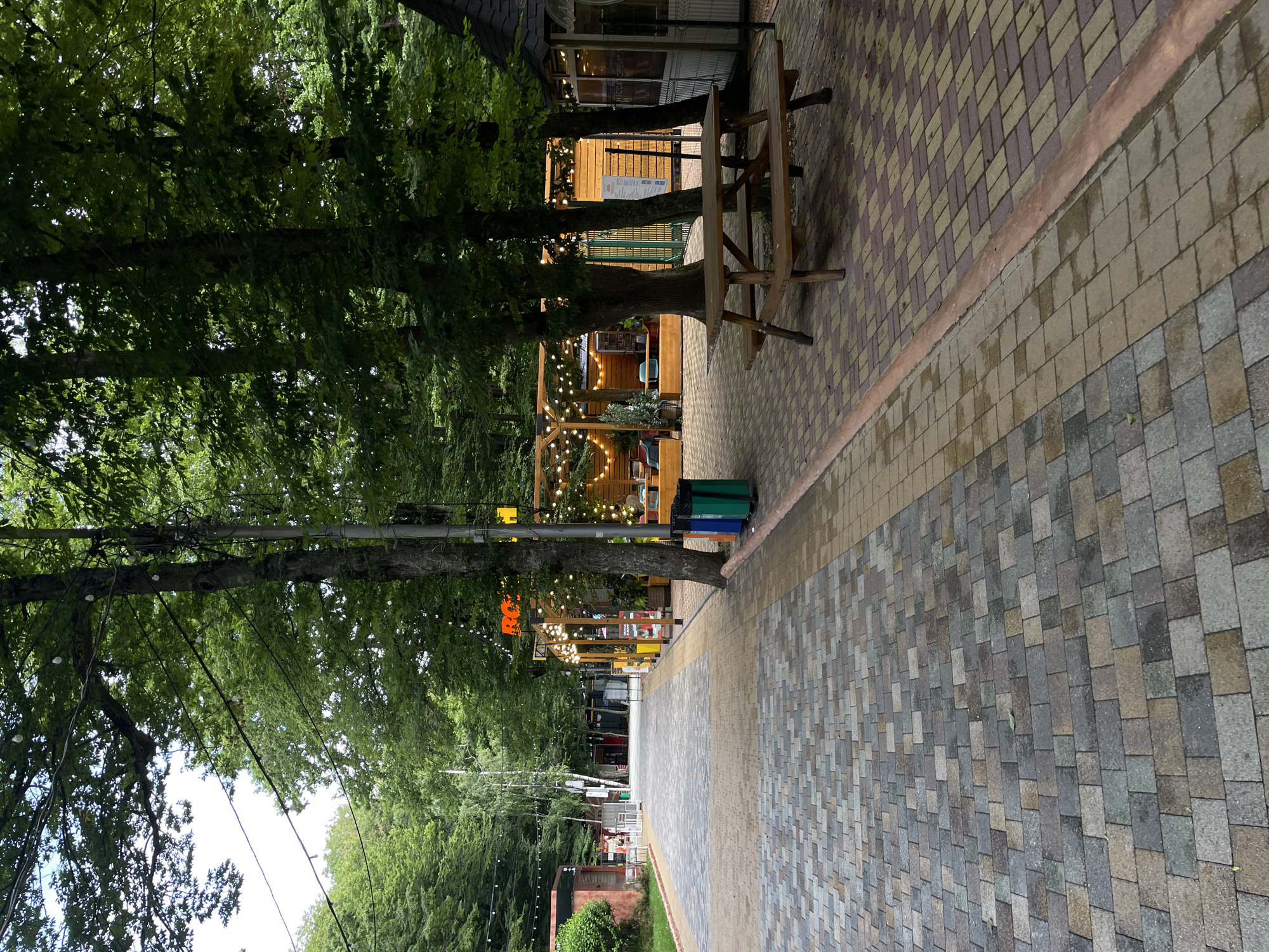 The image depicts an outdoor seating area in a park or recreational space, possibly within a city like Stavropol, Russia. The pathway is paved with multicolored rectangular stones. To the right of the path, a wooden picnic table with benches is positioned. Beyond the table, a small structure with a dark roof and windows is visible. Further down the path, on the left, there are several modular structures with windows, some adorned with outdoor seating and string lights. A prominent sign with orange lettering, possibly a logo or name, is visible above one of these structures. The entire area is shaded by a dense canopy of tall green trees, with some string lights strung between them, suggesting an evening or ambient lighting setup. There are no people clearly visible in the scene. The environment appears to be a well-maintained public space designed for relaxation and outdoor dining or gathering.